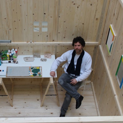 Auf dem Bild ist ein Künstler in einem Atelier aus einem Holzkubus zu sehen.