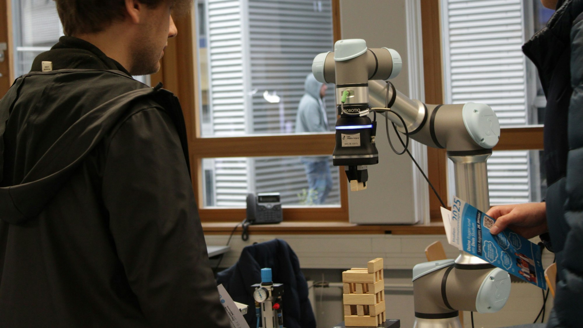 Ein Roboter greift einen Holzklotz und legt ihn auf einen Stapel mit Klötzen, zwei Besucher schauen zu.