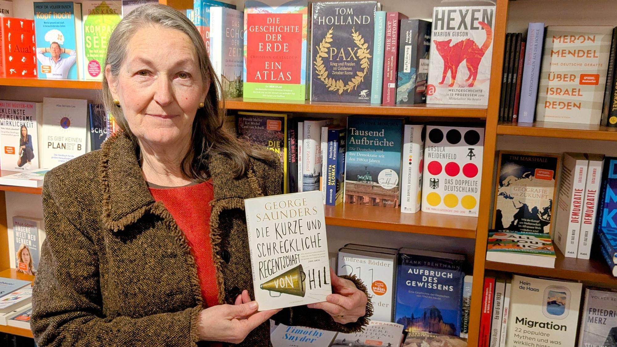Iris Pütz steht vor einem Bücherregal und präsentiert das Buch „Die kurze und schreckliche Regentschaft von Phil“ von George Saunders.