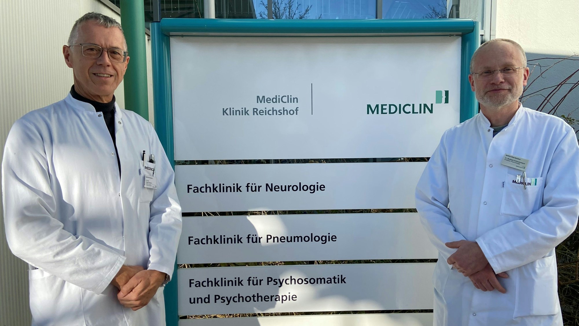 Matthias Schmalenbach, Chefarzt der Pneumologie an der Mediclin-Rehaklinik in Reichshof-Eckenhagen, und Jürgen Bonnert, Chefarzt der Neurologie und kommissarischer Chefarzt für Psychosomatik und Psychotherapie, stehen vor einem Klinikschild.