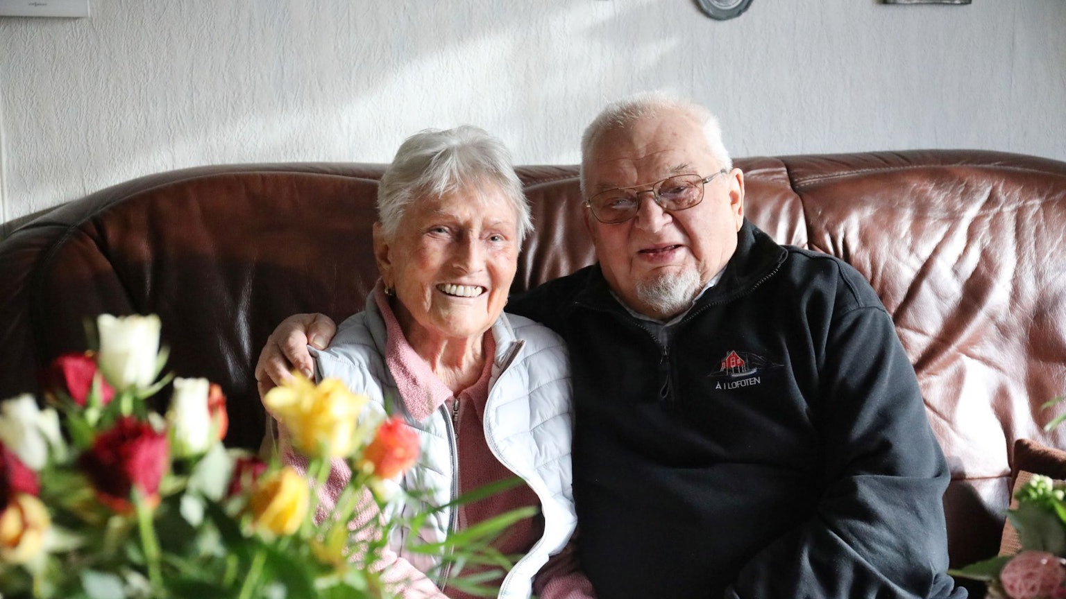 Ein Ehepaar sitzt nebeneinander auf einem Sofa, am Rand steht ein bunter Blumenstrauß.