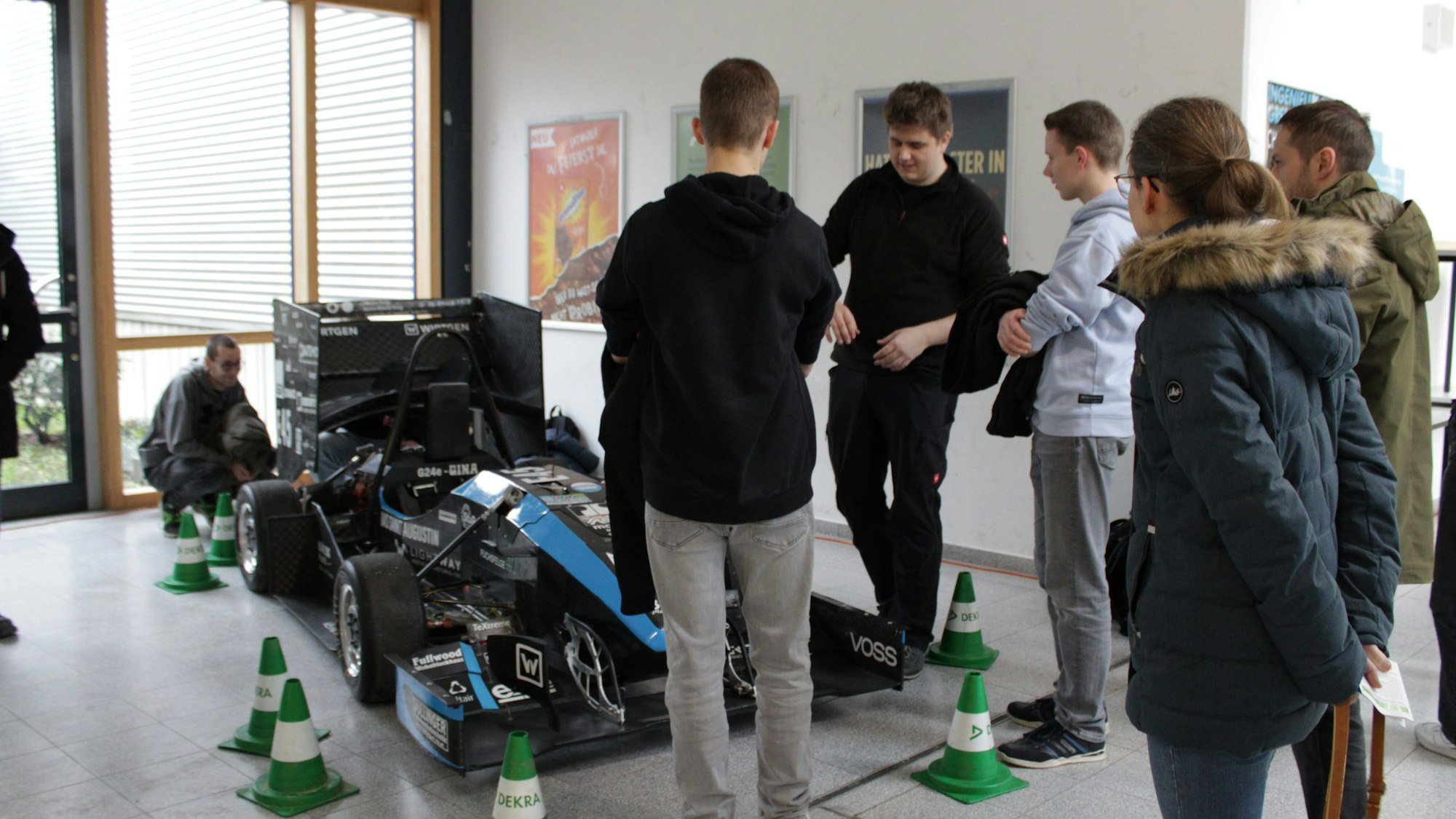 Mehrere Schülerinnen und Schüler stehen vor einem Rennwagen im Flur einer Hochschule, ein Student steht daneben und erzählt etwas.