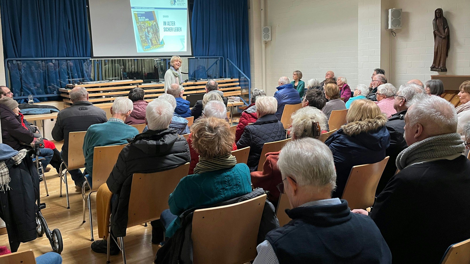 Eine Frau spricht vor einer Gruppe von Senioren in einem Pfarrheim.
