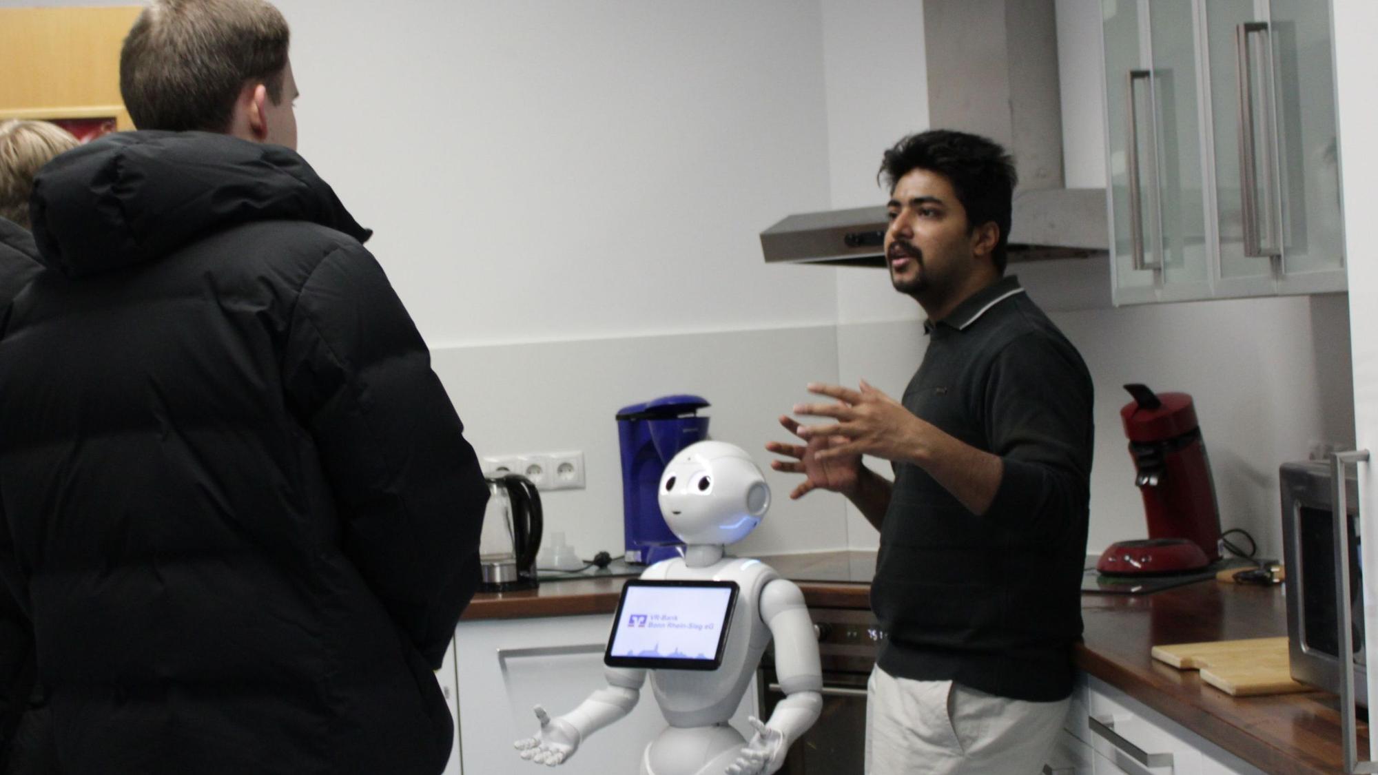 Ein Student steht neben einem Roboter, der wie er mit den Händen gestikuliert, und spricht mit zwei Schülern.