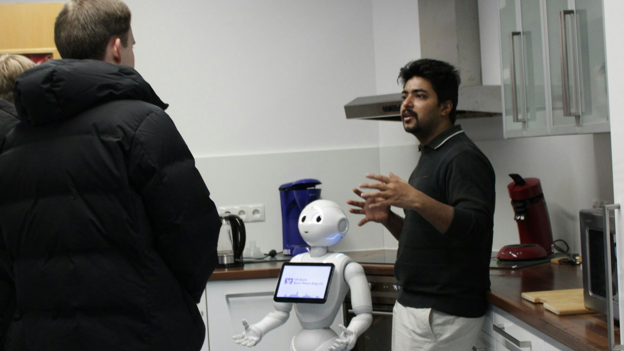 Ein Student steht neben einem Roboter, der wie er mit den Händen gestikuliert, und spricht mit zwei Schülern.