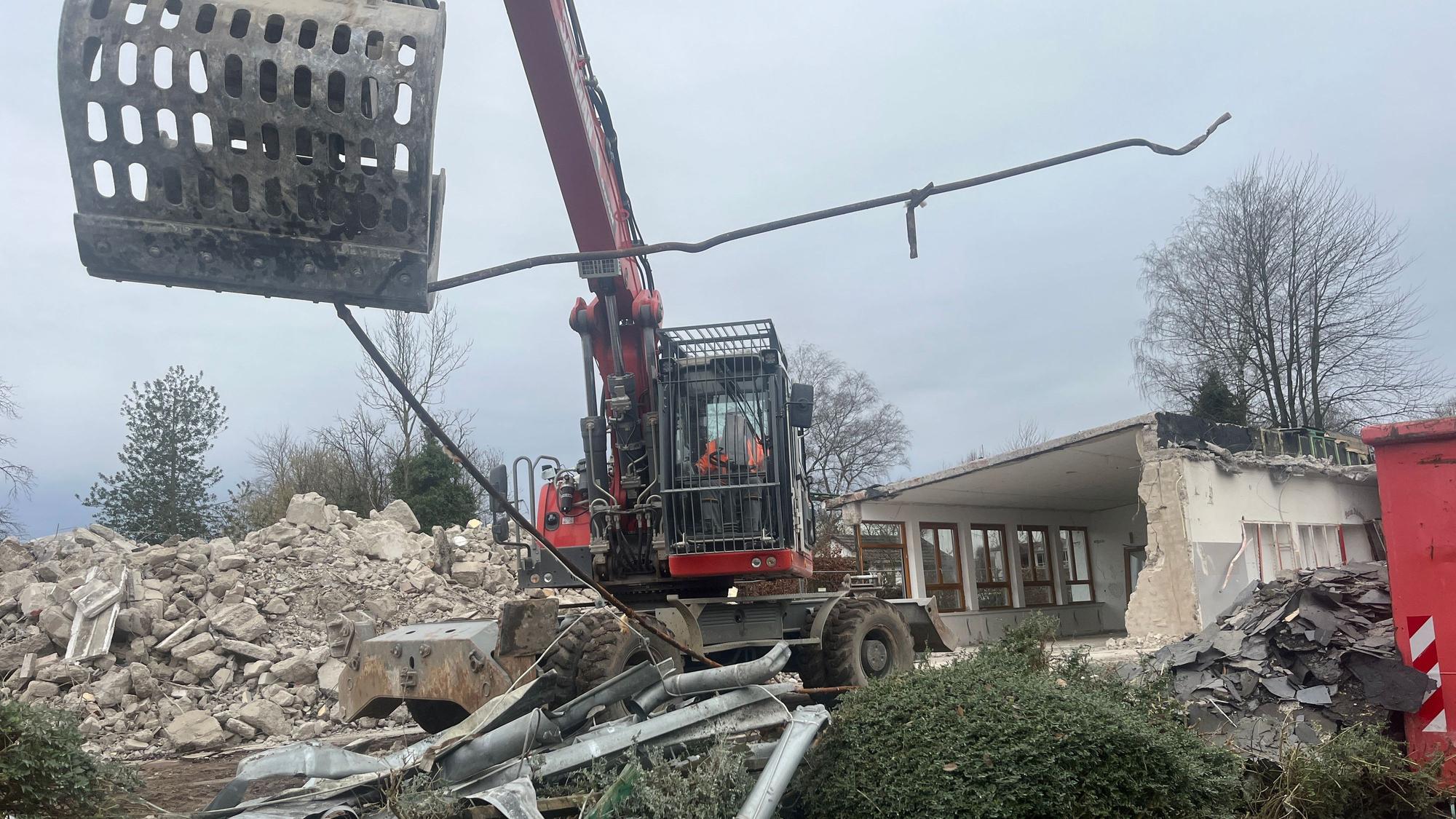 Ein Bagger steht vor einem abgerissenen Gebäude und biegt Metall.