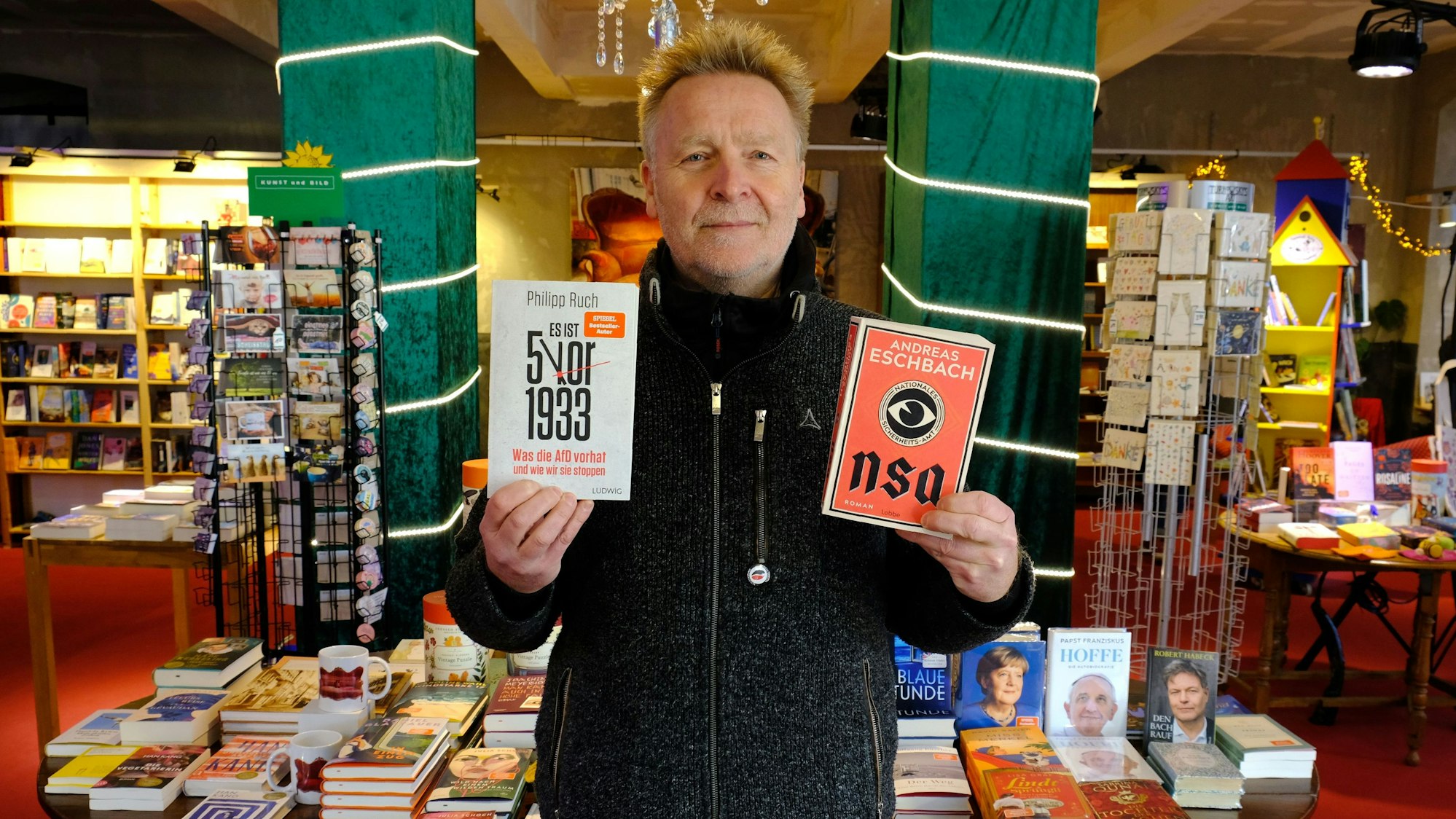 Josef Mütter steht mit zwei Büchern in den Händen in seiner Buchhandlung.