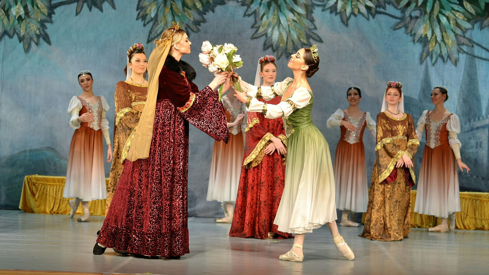 Eine Ballett-Tänzerin in grün-weißem Kleid überreicht einer anderen Tänzerin in dunkelroter Robe einen Strauß Blumen. Im Hintergrund sind weitere Tänzerinnen in prachtvollen Kostümen zu sehen.