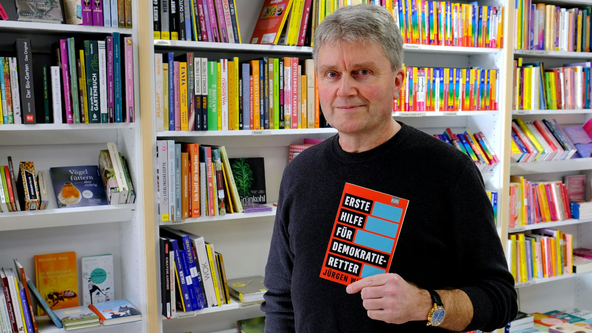 Buchhändler Thomas Pavlik steht mit dem Buch „Erste Hilfe für Demokratie-Retter“ vor einem Bücherregal.