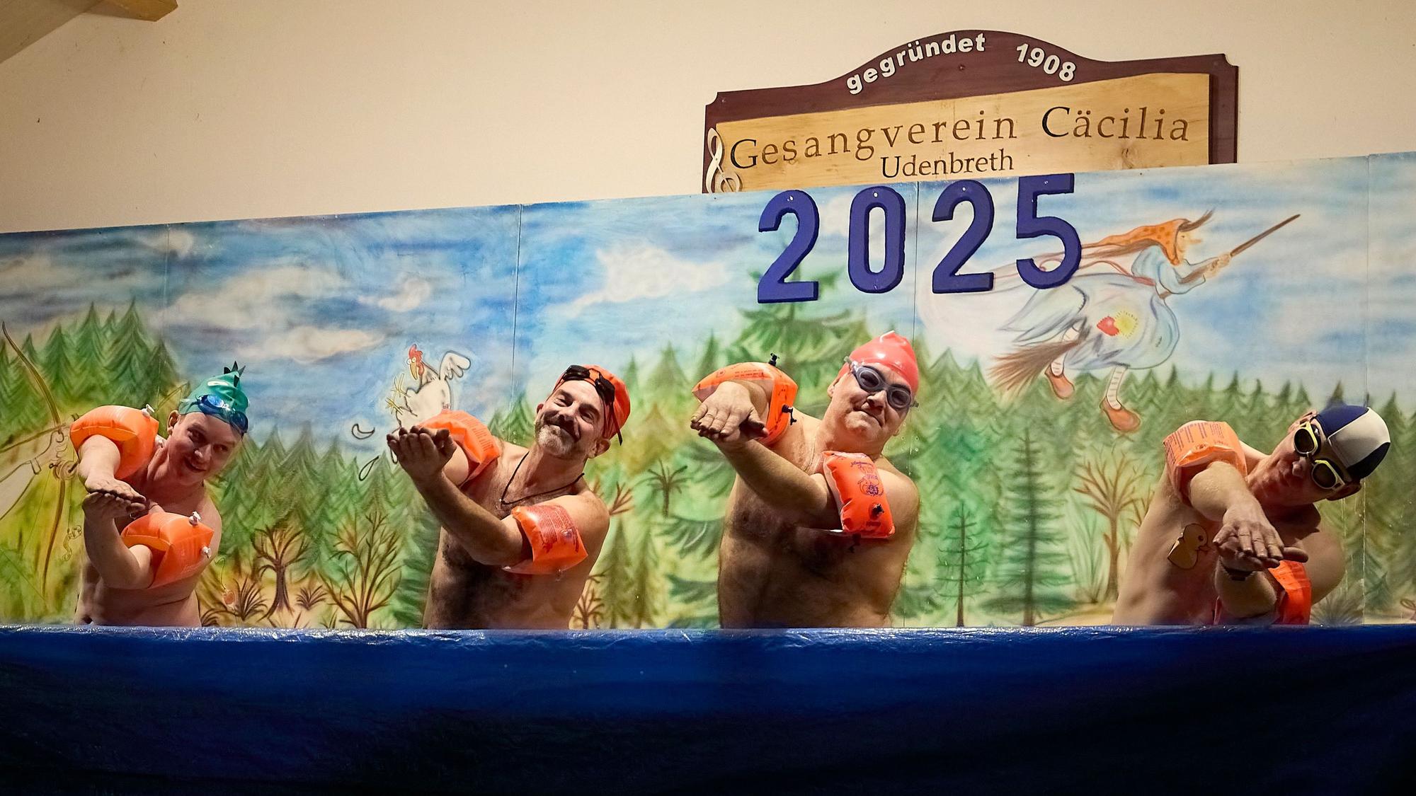 Vier Männer mit bloßem Oberkörper und Schwimmflügeln zeigen vor einem Bühnenbild Bewegungen wie beim Synchronschwimmen.