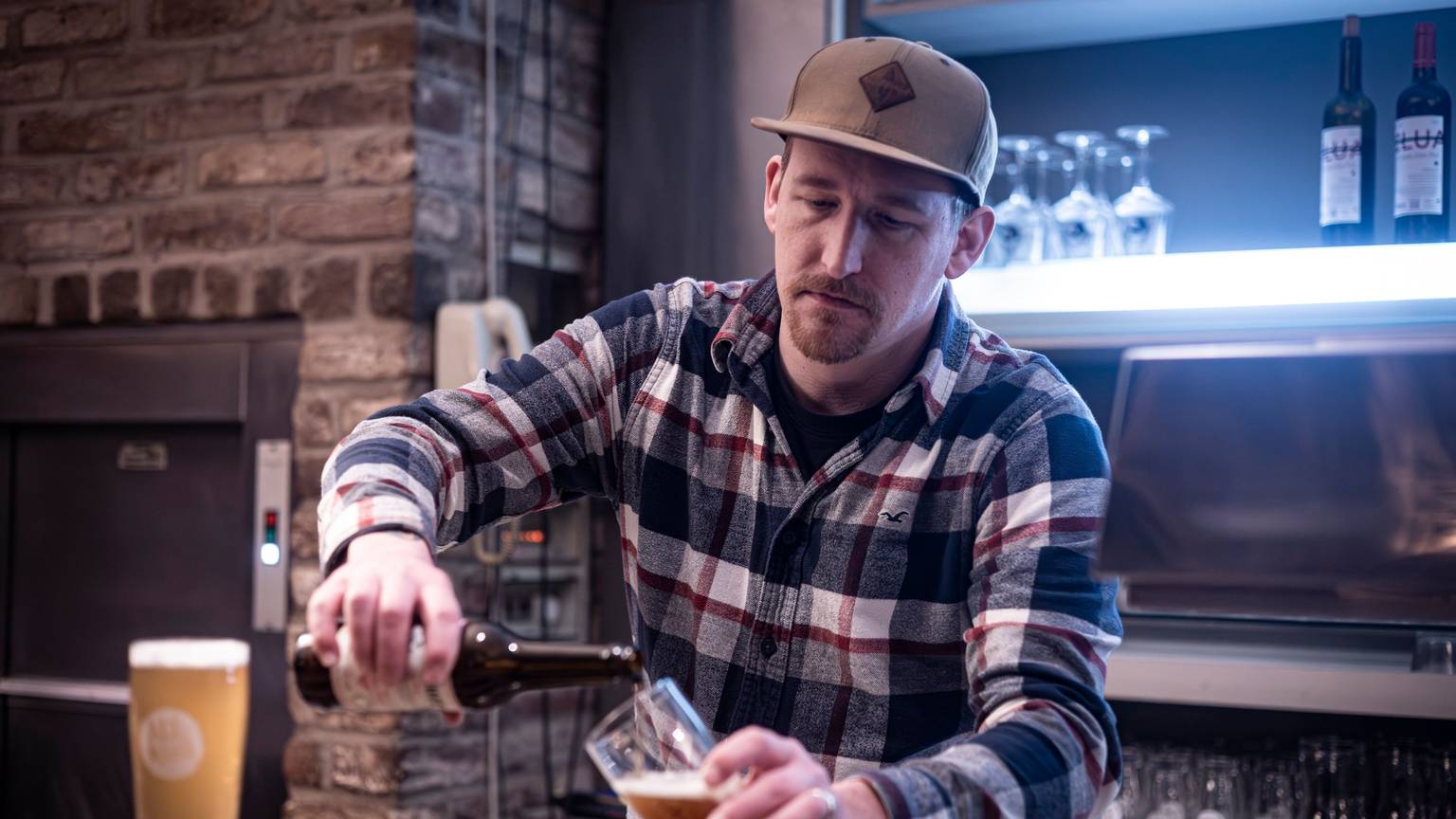 Balthasar Fischer hinter der Theke, er schenkt Bier aus einer Flasche ins Glas.