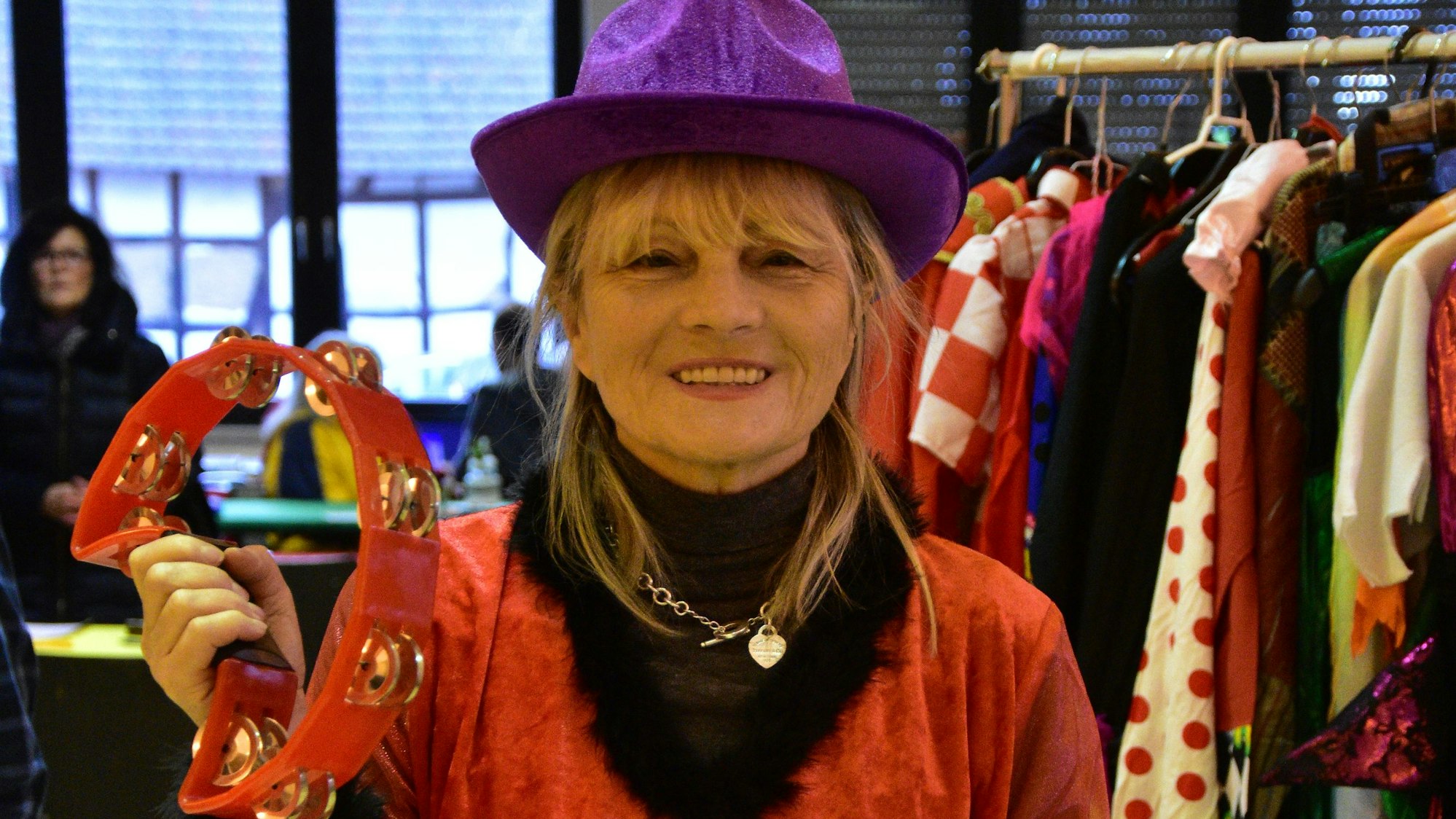 Eine lächelnde Frau trägt ein rotes Samt-Gewand und einen lila Hut, in der Hand einen Glockenkranz.