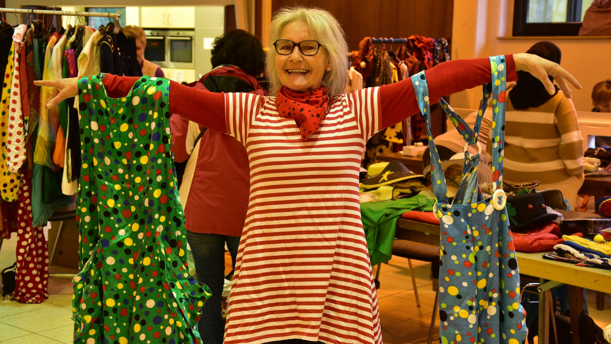 Eine Frau in geringeltem Kleid hält breit lächelnd ein Clown-Kostüme an jedem Arm.