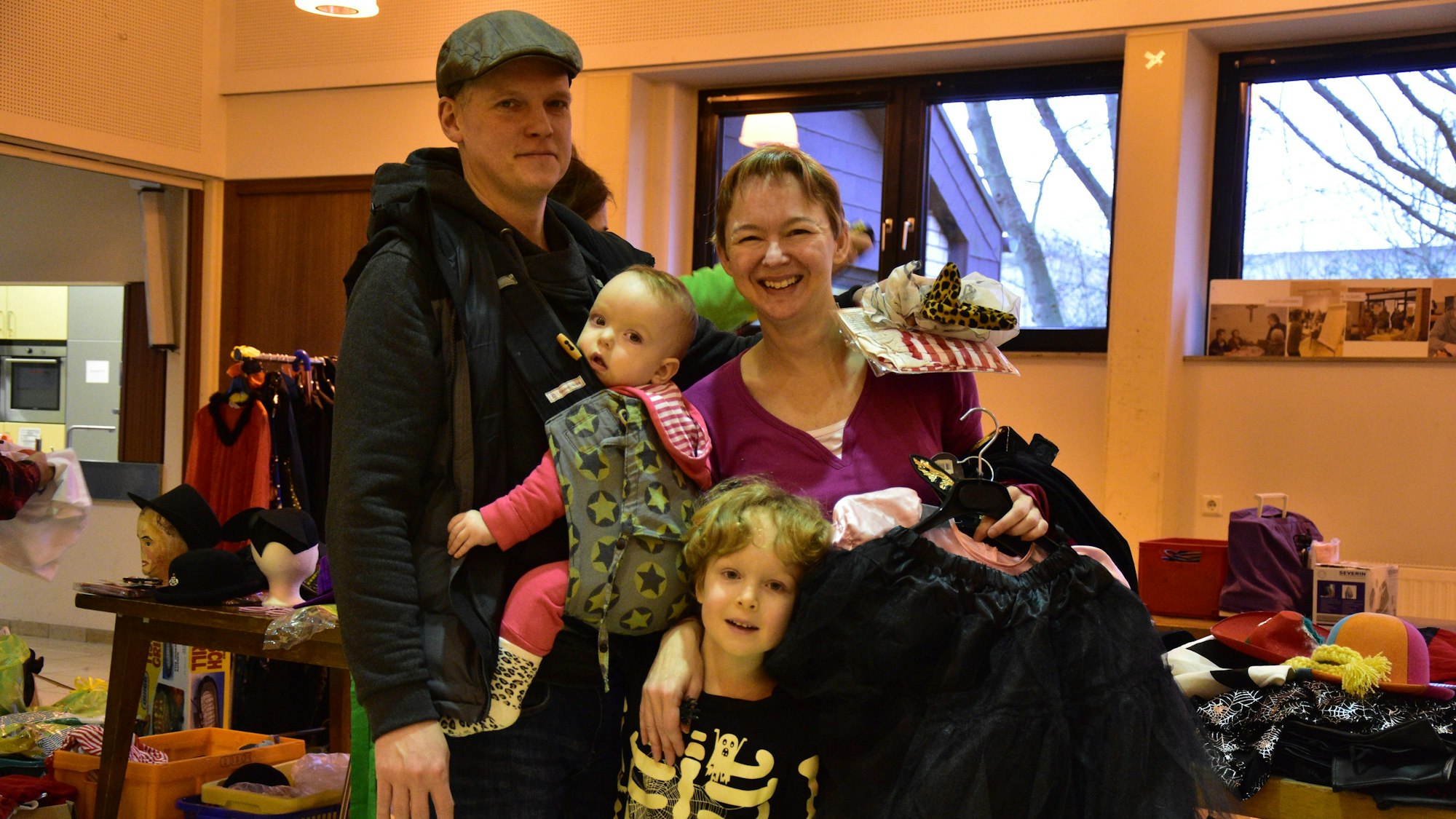 Eine Familie mit zwei Kindern steht in einem Raum, in dem einige Kostüme und Accessoires zu sehen sind. Der Junge trägt ein Skelettkostüm, die Mutter hält mehrere Bügel mit Kleidung in der Hand.