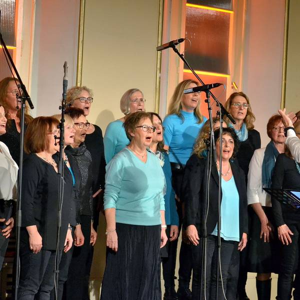 Frauen stehen auf einer Bühne und singen, den Dirigenten sieht man von hinten.
