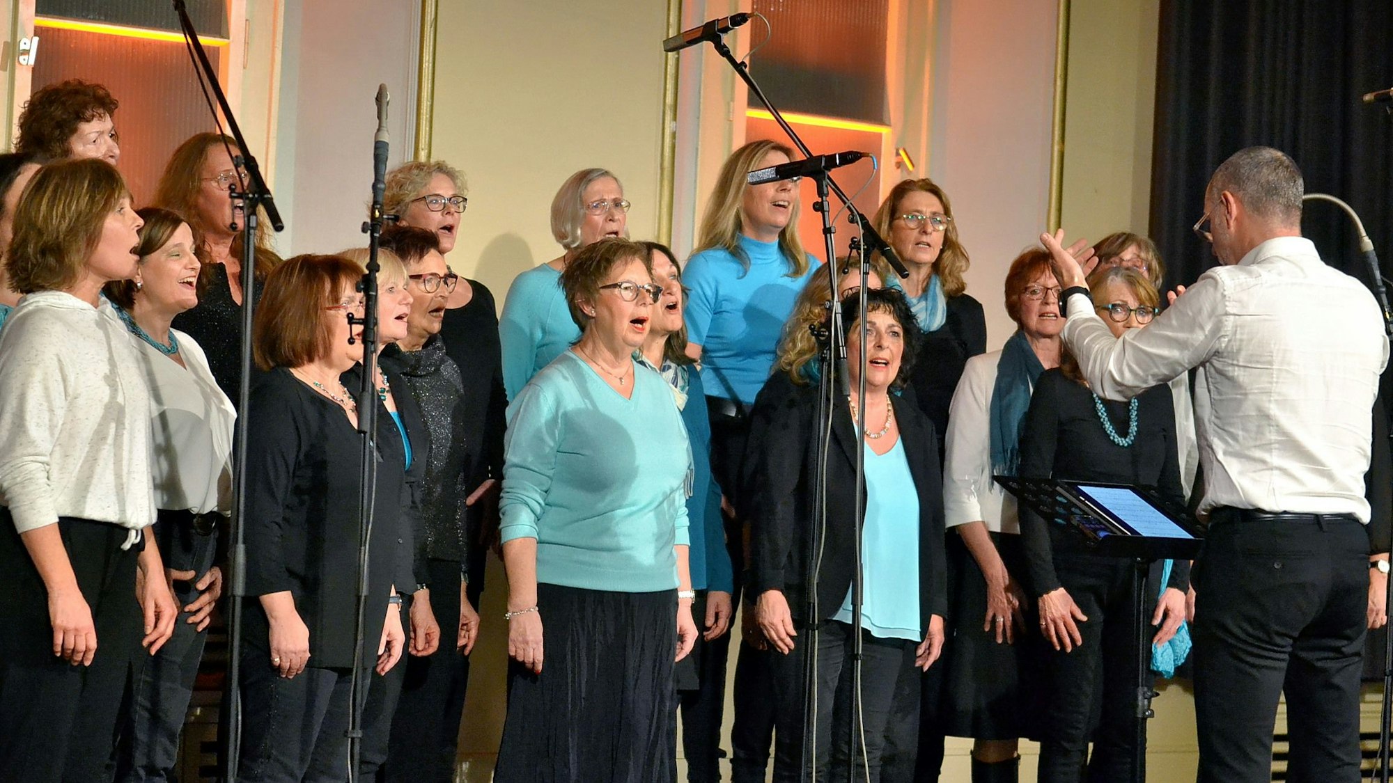 Frauen stehen auf einer Bühne und singen, den Dirigenten sieht man von hinten.