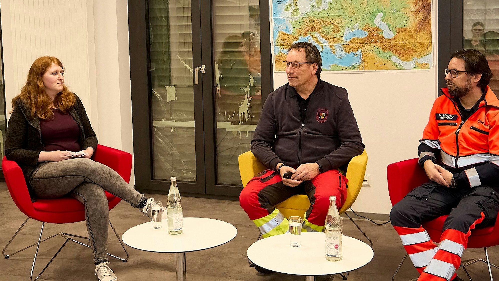 Zwei Männer und eine Frau sitzen in Sesseln an zwei weißen Tischen im Haus der Begegnung in Kall.