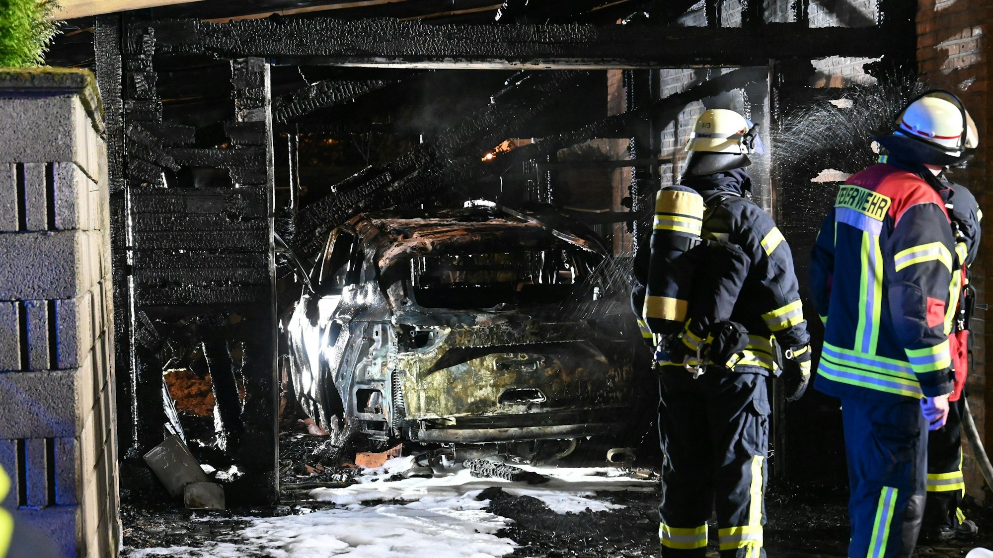 Eine abgebrannte Garage mit einem zerstörten Auto. Am Auto und auf dem Boden sind noch Reste des Löschschaums zu sehen.