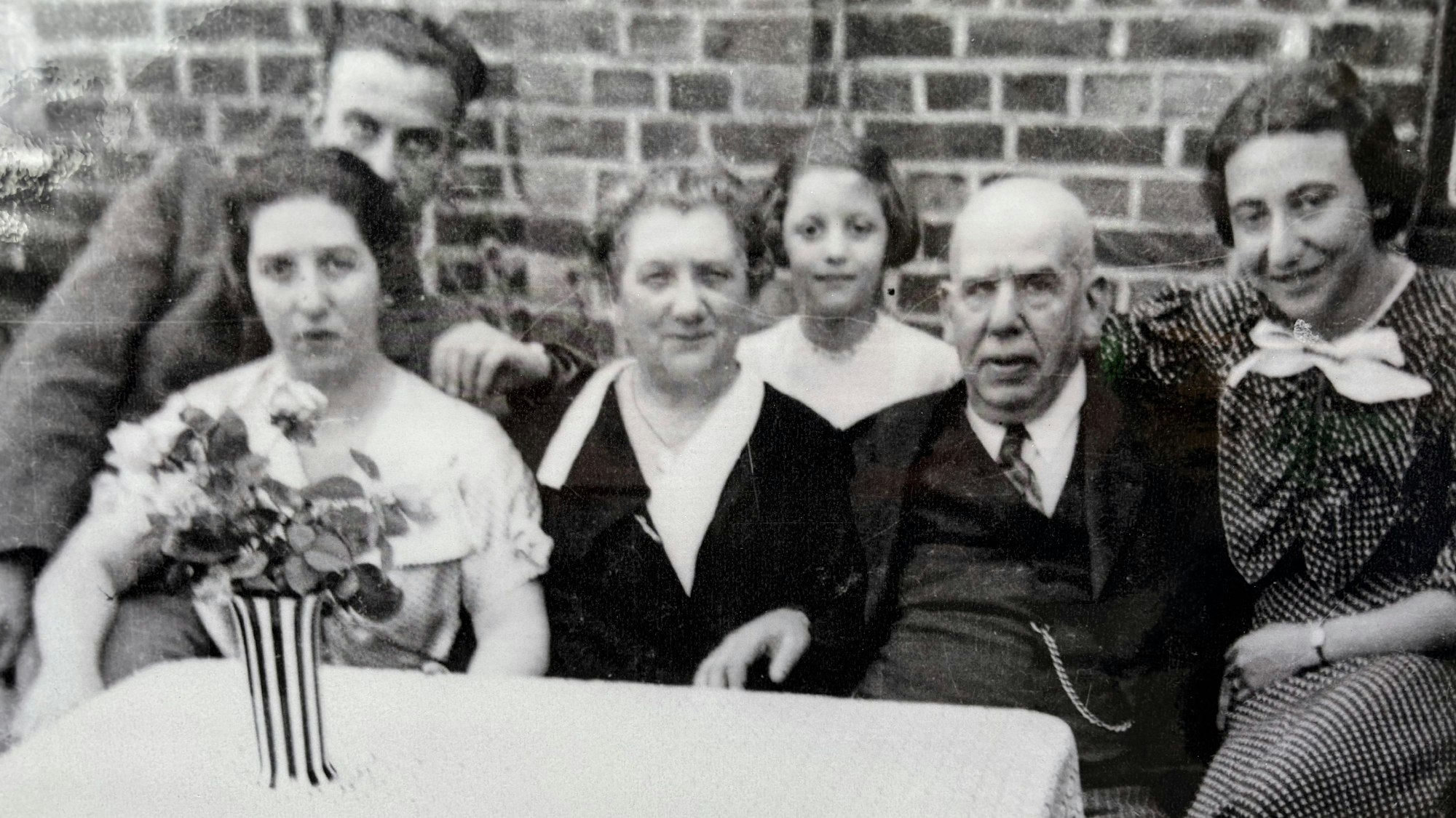 Eine Schwarz-Weiß-Aufnahme zeigt mehrere Menschen, die draußen an einem Tisch sitzen, auf dem eine Vase mit Blumen steht.