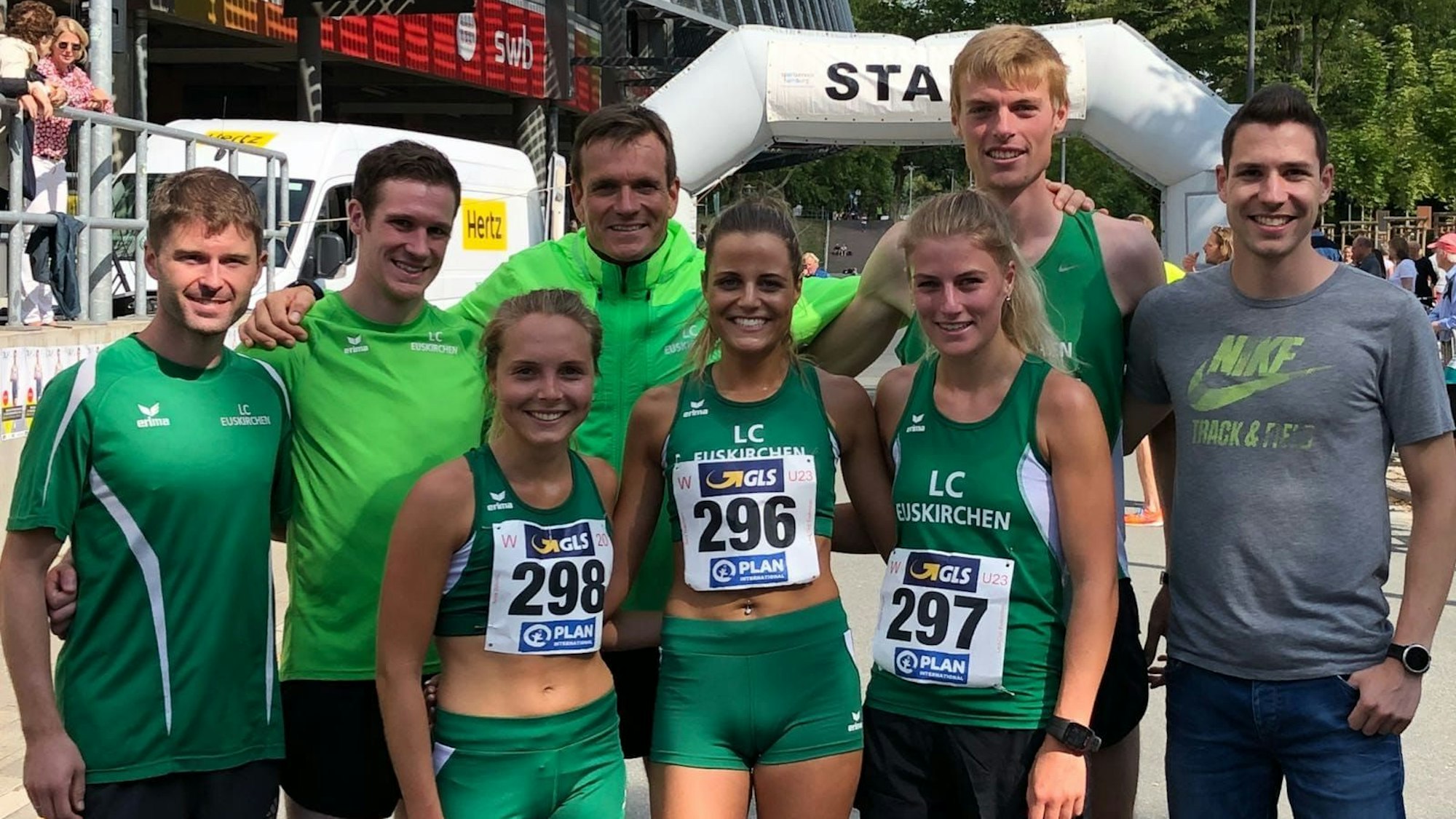 Drei Frauen im Läuferdress stehen Arm in Arm vorne, dahinter fünf Männer, ebenfalls größtenteils in Sportkleidung und Arm in Arm. Im Hintergrund ist ein Starttor zu sehen.