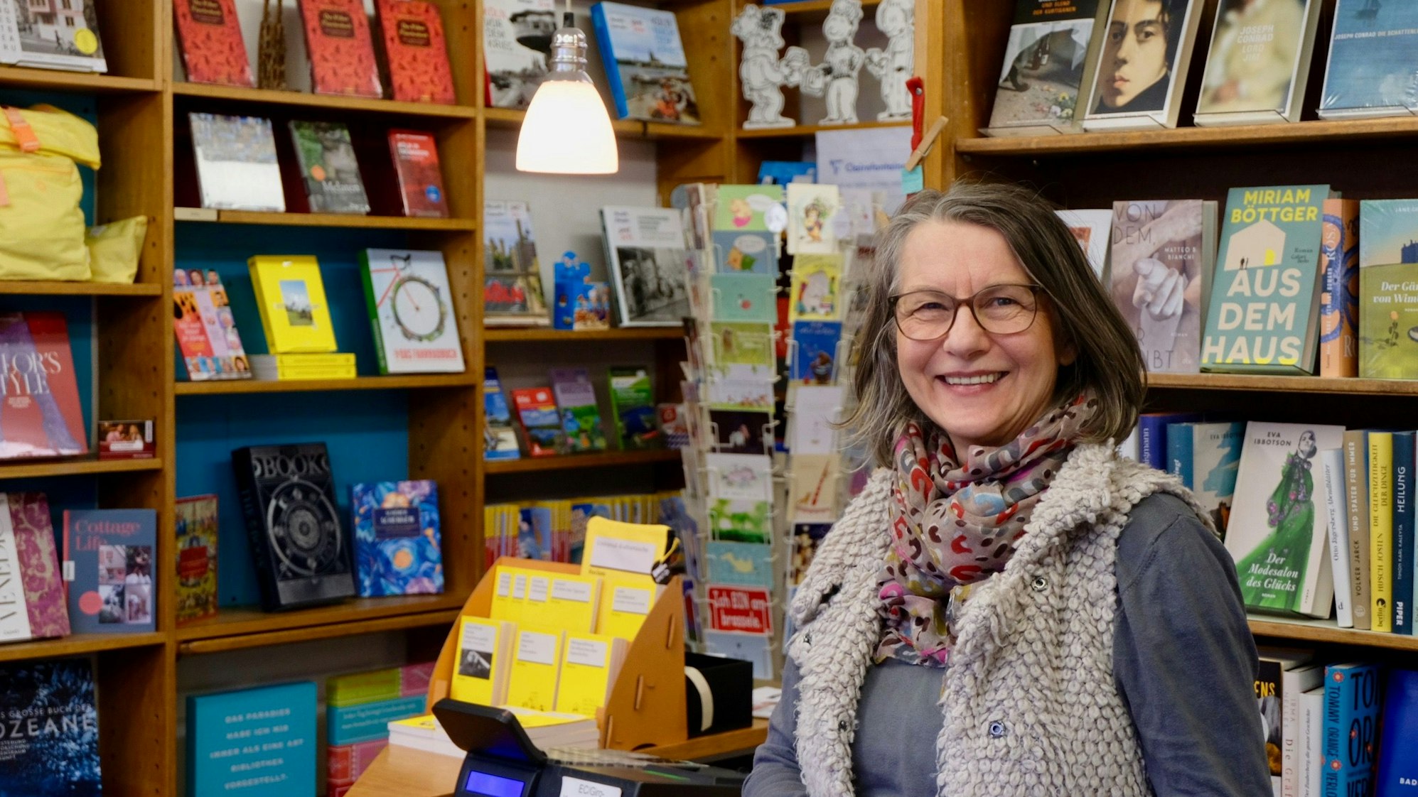 Eine Frau mit Brille, schulterlangem Haar und buntem Schal um den Hals steht lachend vor einem Bücherregal.