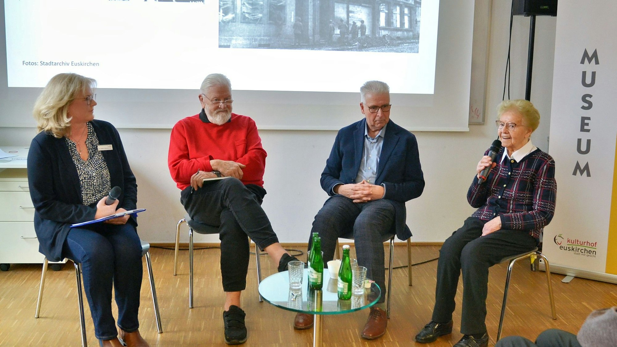 Zwei Frauen und zwei Männer sitzen nebeneinander auf Stühlen, vor ihnen ein Glastisch mit Getränken.