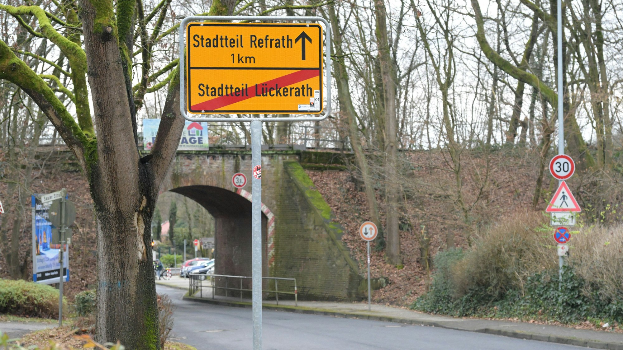 Ein gelbes Ortsschild mit dem Schriftzug „Stadtteil Refrath“ steht vor einer Unterführung.