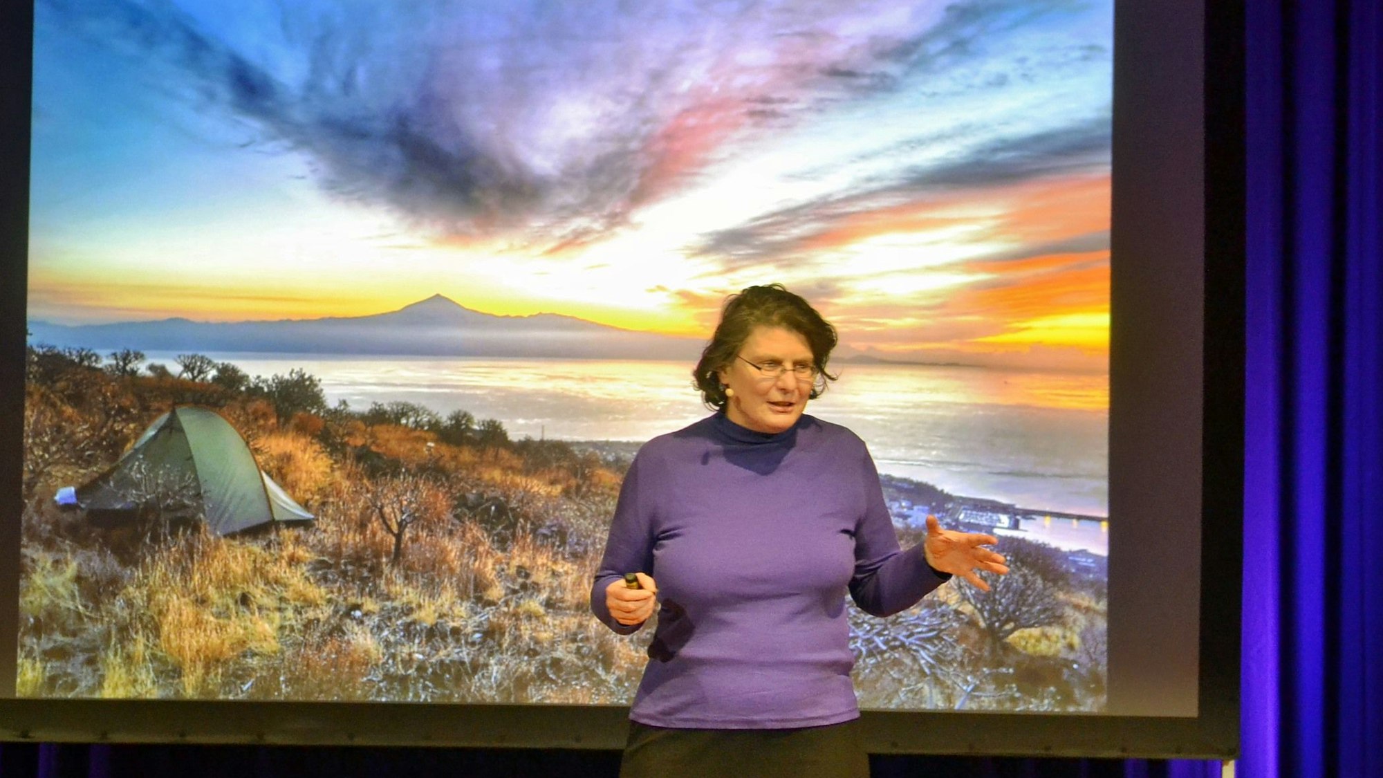 Christine Thürmer steht vor einer Leinwand, auf die ein Bild projiziert wird. Auf diesem sind ein Sonnenuntergang zu sehen und im Vordergrund ein Zelt.