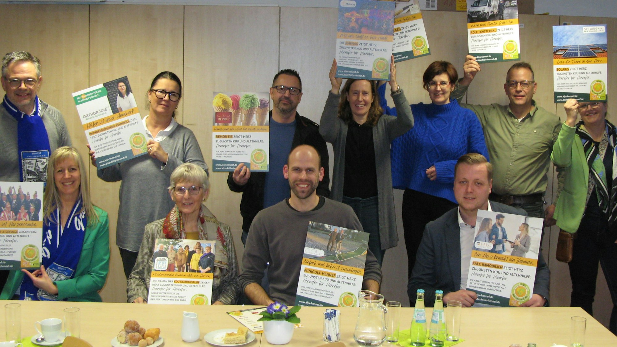 Zwei Frauen und zwei Männer sitzen nebeneinander an einem Tisch. Dahinter stehen vier Frauen und drei Männer. Alle halten Plakate hoch.