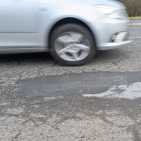 Das Foto zeigt das reparierte Schlagloch im Asphalt der Luxemburger Straße, dahinter ein vorbeifahrendes Auto.