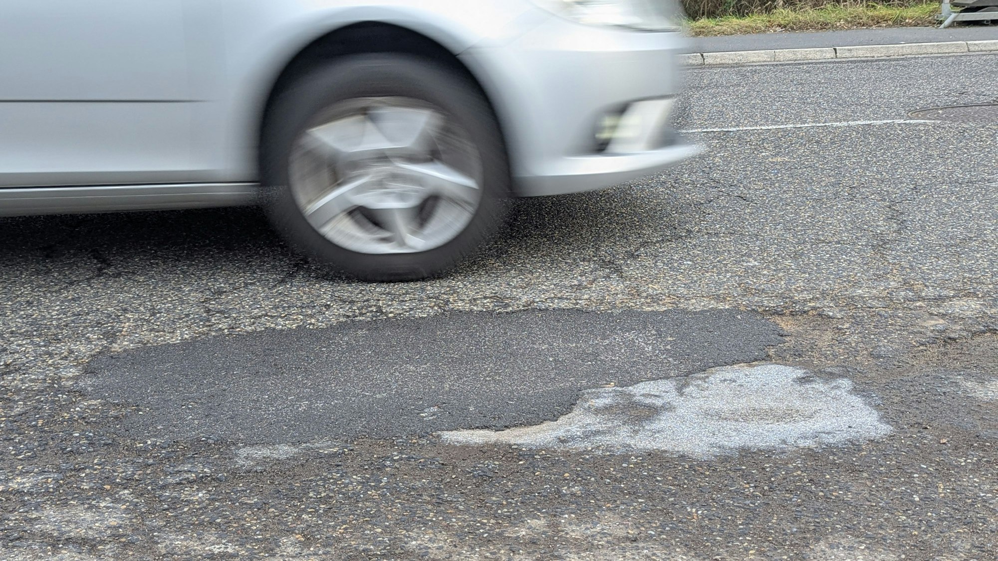 Das Foto zeigt das reparierte Schlagloch im Asphalt der Luxemburger Straße, dahinter ein vorbeifahrendes Auto.