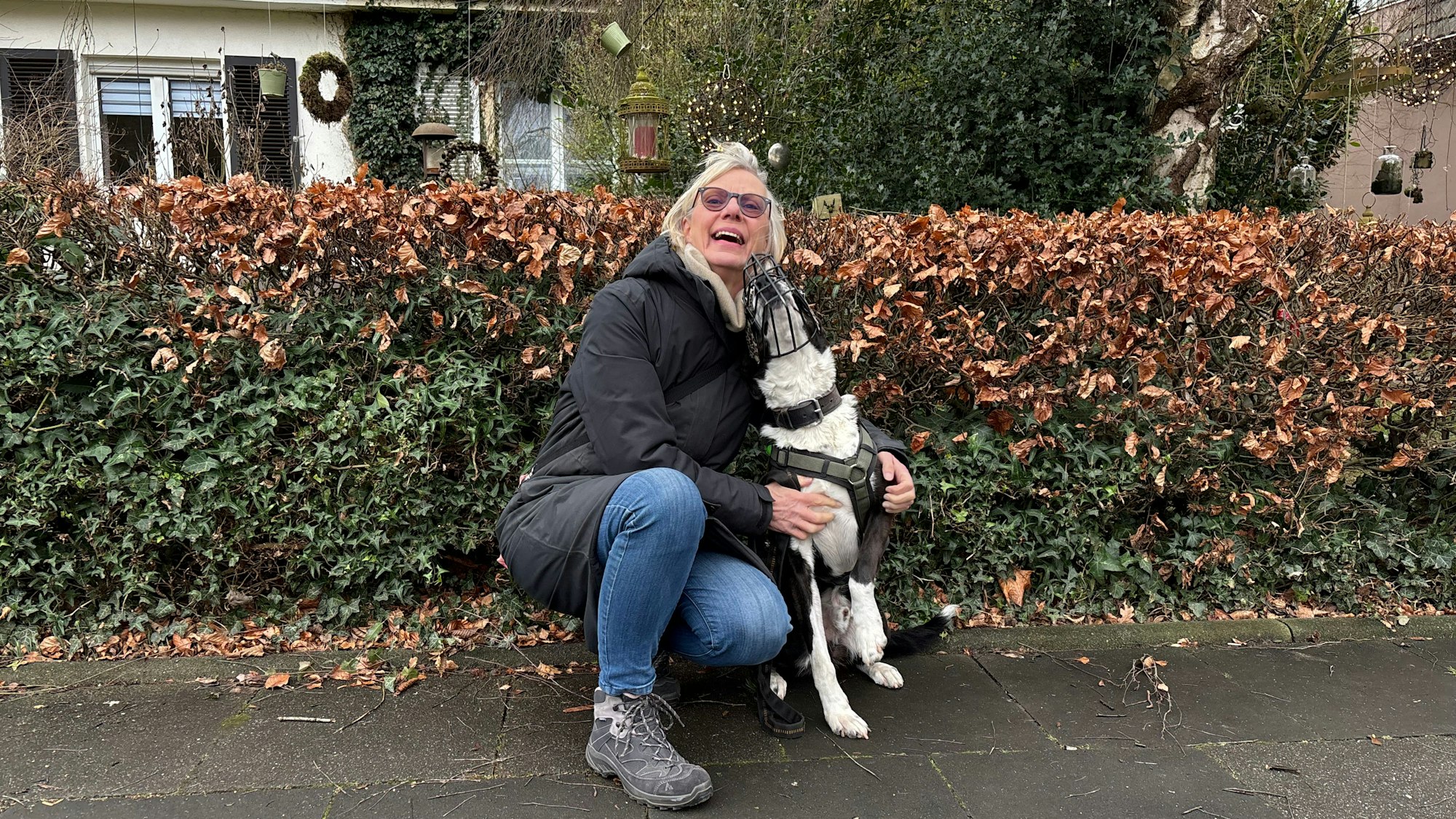 Eine ältere Frau kniet neben einem Hund. Der Mischling trägt einen Maulkorb. Die Frau lacht.
