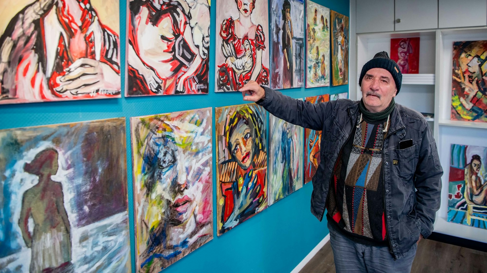 Das Bild zeigt den Künstler, der mit seinem rechten Arm auf einige seiner Bilder zeigt.