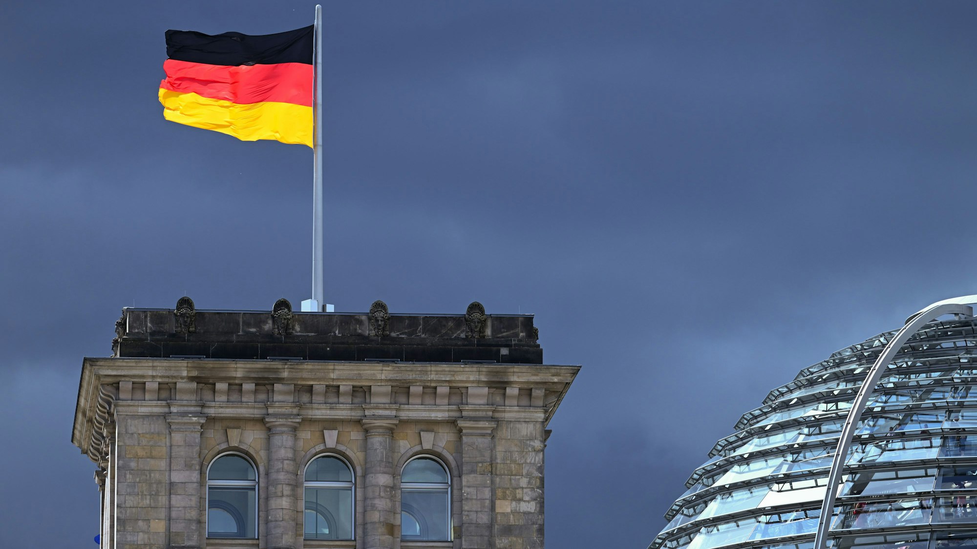 ARCHIV - 03.08.2023, Berlin: Eine der Flaggen auf dem Reichstag leuchtet bei starkem Wind wehend vor der Kulisse von dunklen Regenwolken.
