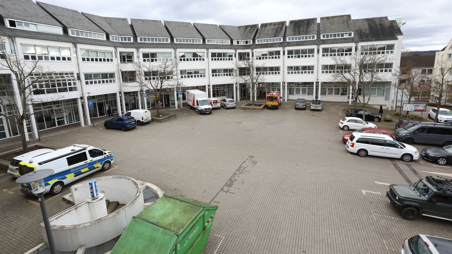 Ein Gebäude mit einer großen gepflasterten Fläche davor, auf der zum Teil Autos stehen.