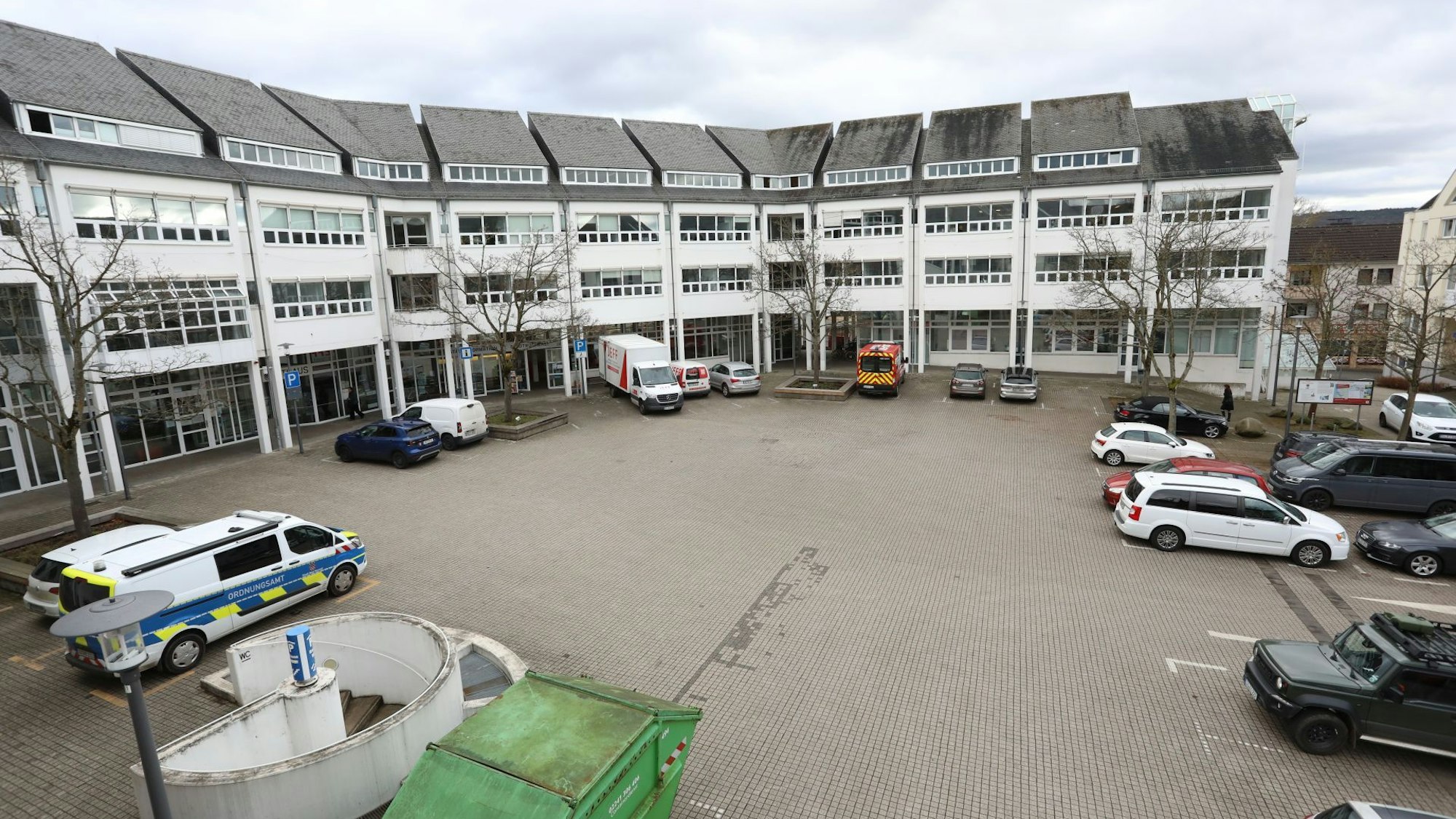 Ein Gebäude mit einer großen gepflasterten Fläche davor, auf der zum Teil Autos stehen.