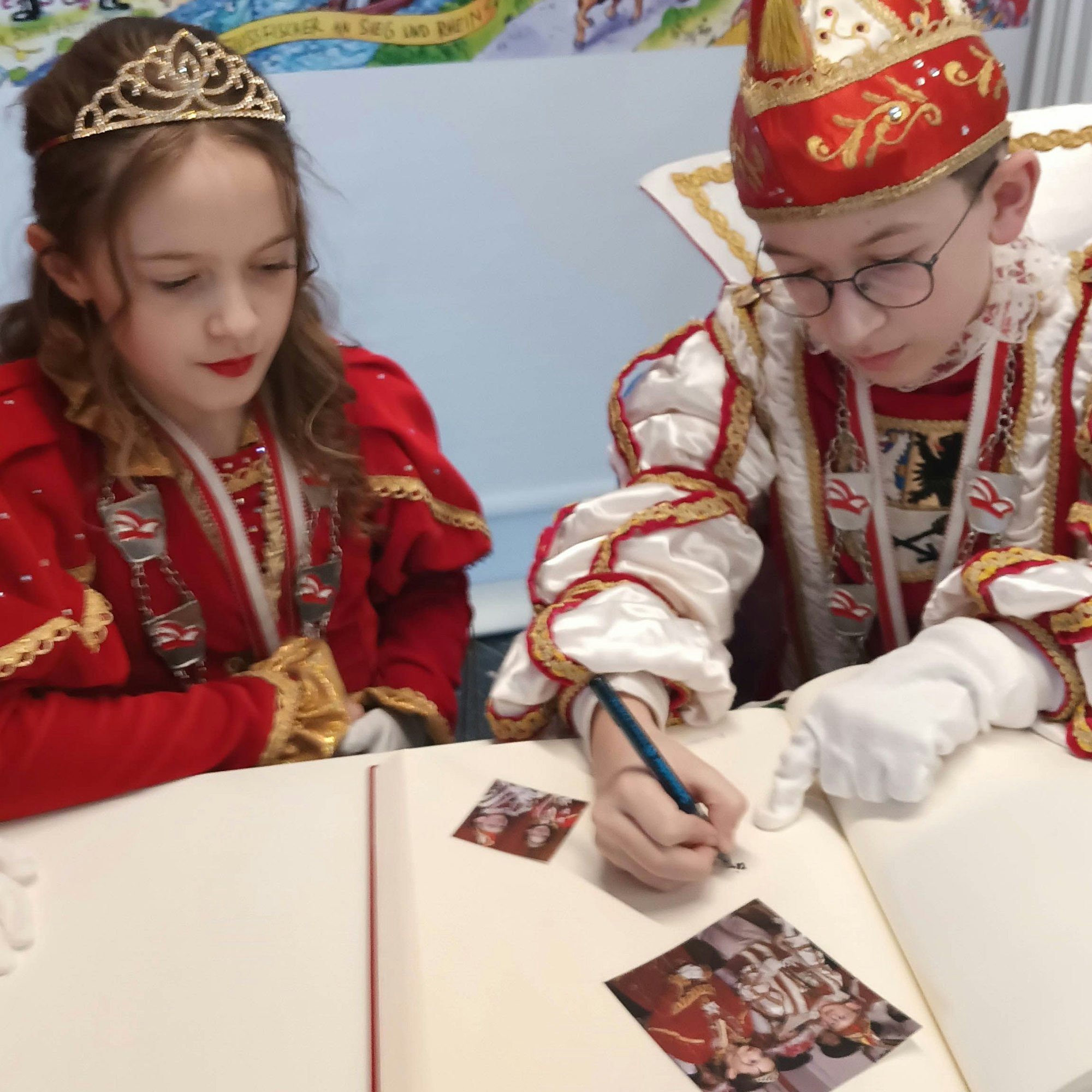 Das Kaller Kinderprinzenpaar sitzt in seinen Kostümen an einem Tisch. Sie haben Bilder in ein Buch geklebt, nun schreibt der Prinz einen Text dazu.