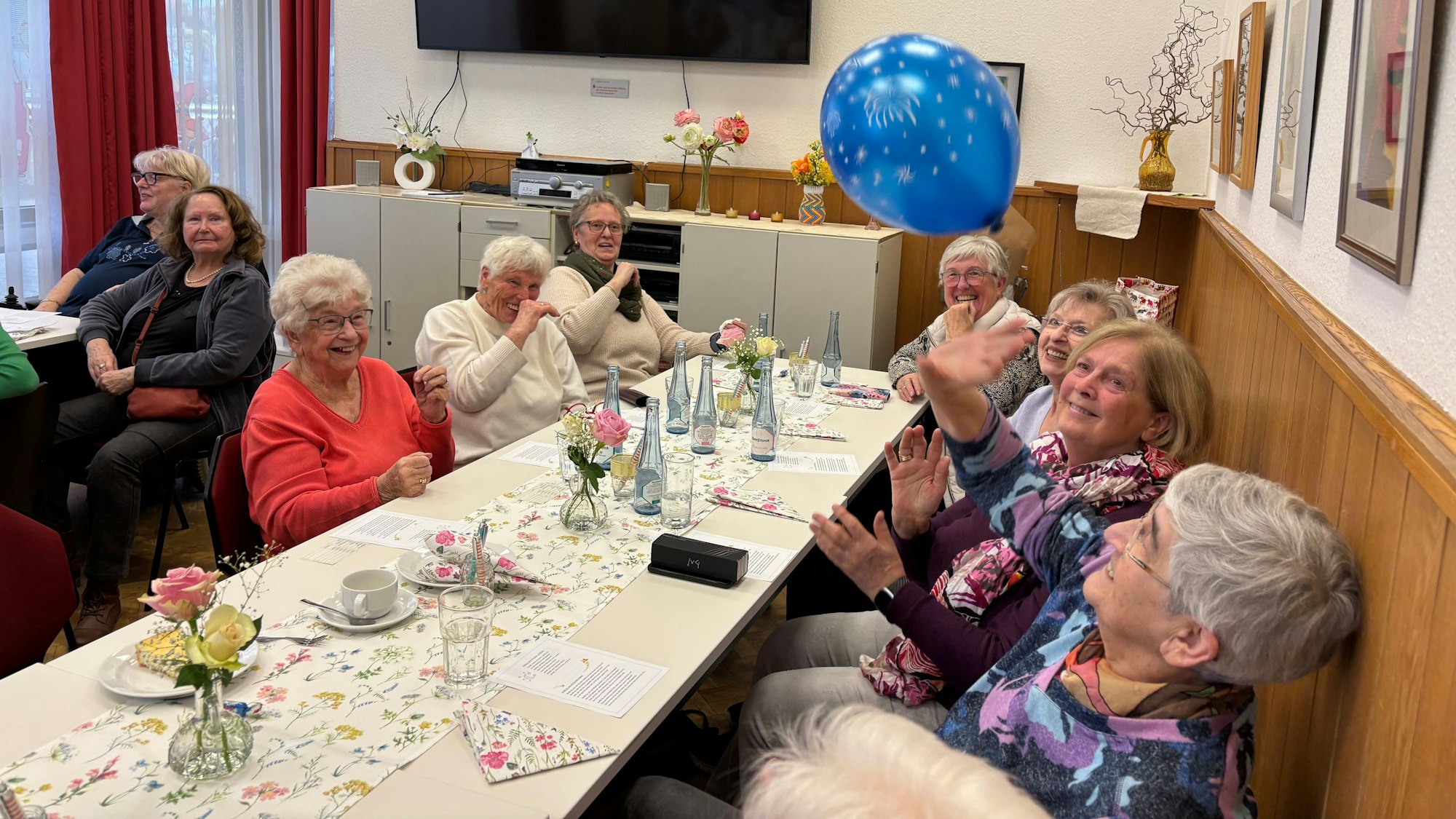 Eine Gruppe älterer Frauen, an einem gedeckten Tisch, spielt sich einen Luftballon zu.
