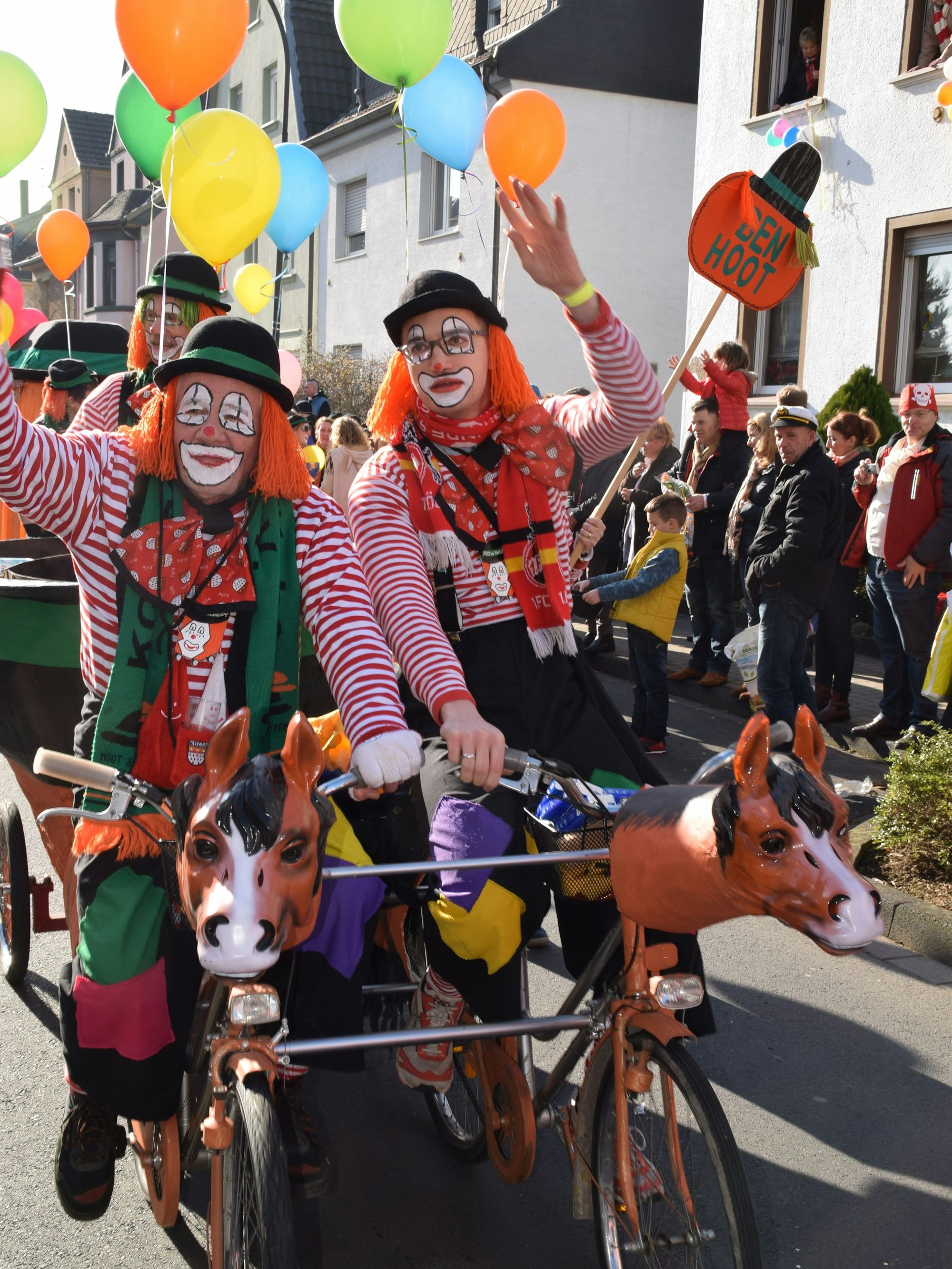 Männer in Clownskostümen fahren auf einem Doppelfahrrad. Am Straßenrand stehen Zuschauer.