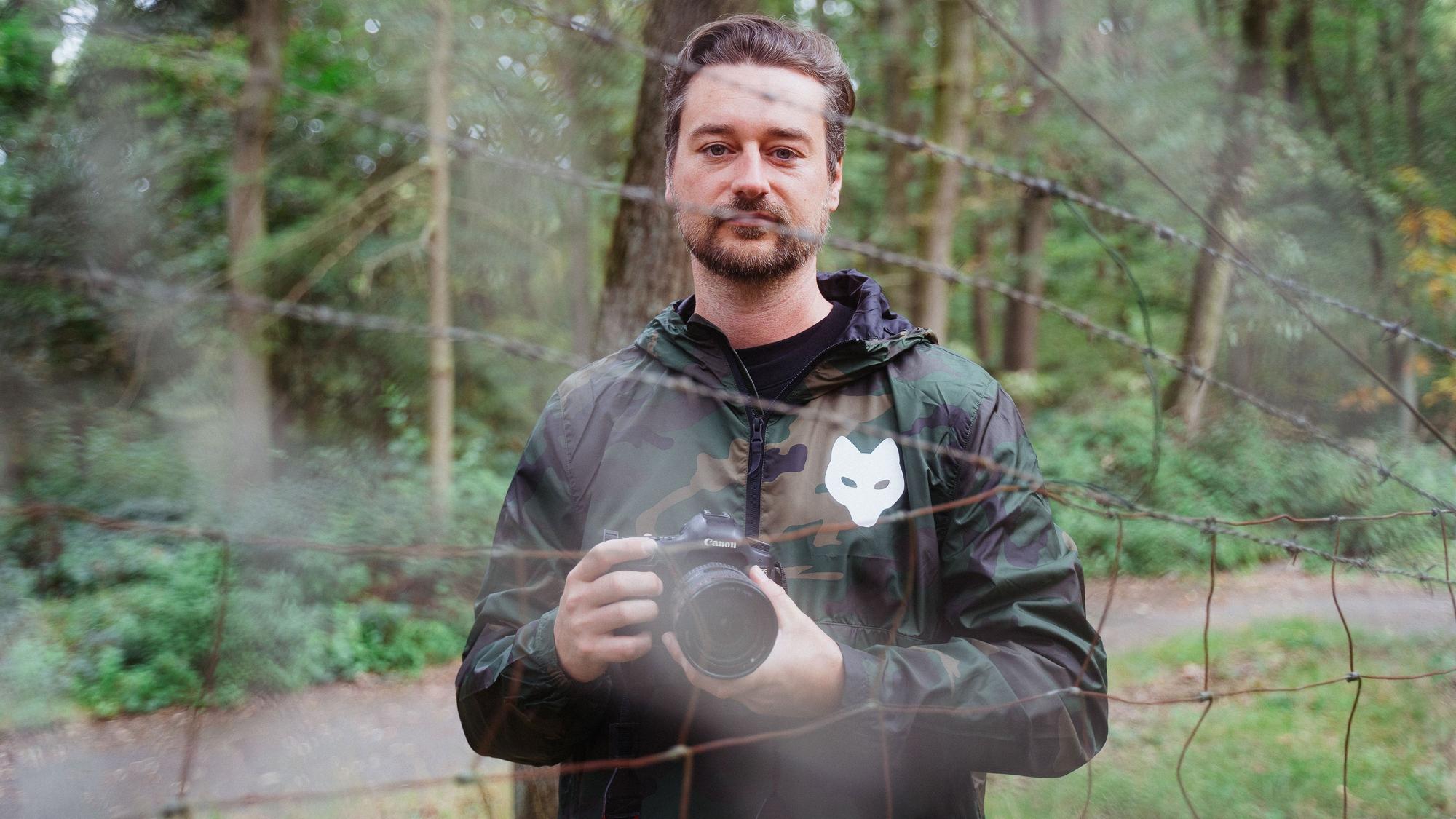 Ein Mann mit einer Kamera steht vor Bäumen und hinter einem Stacheldrahtzaun.