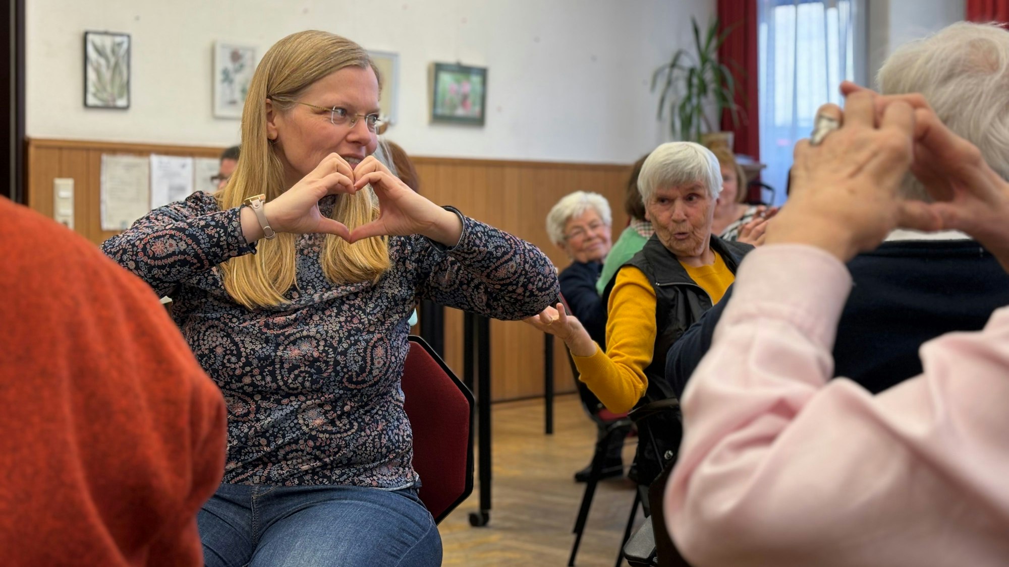 Eine sitzende Frau mit Brille formt ein Herz mit ihren Händen. Ältere Frauen sitzen um sie herum und bewegen sich mit.