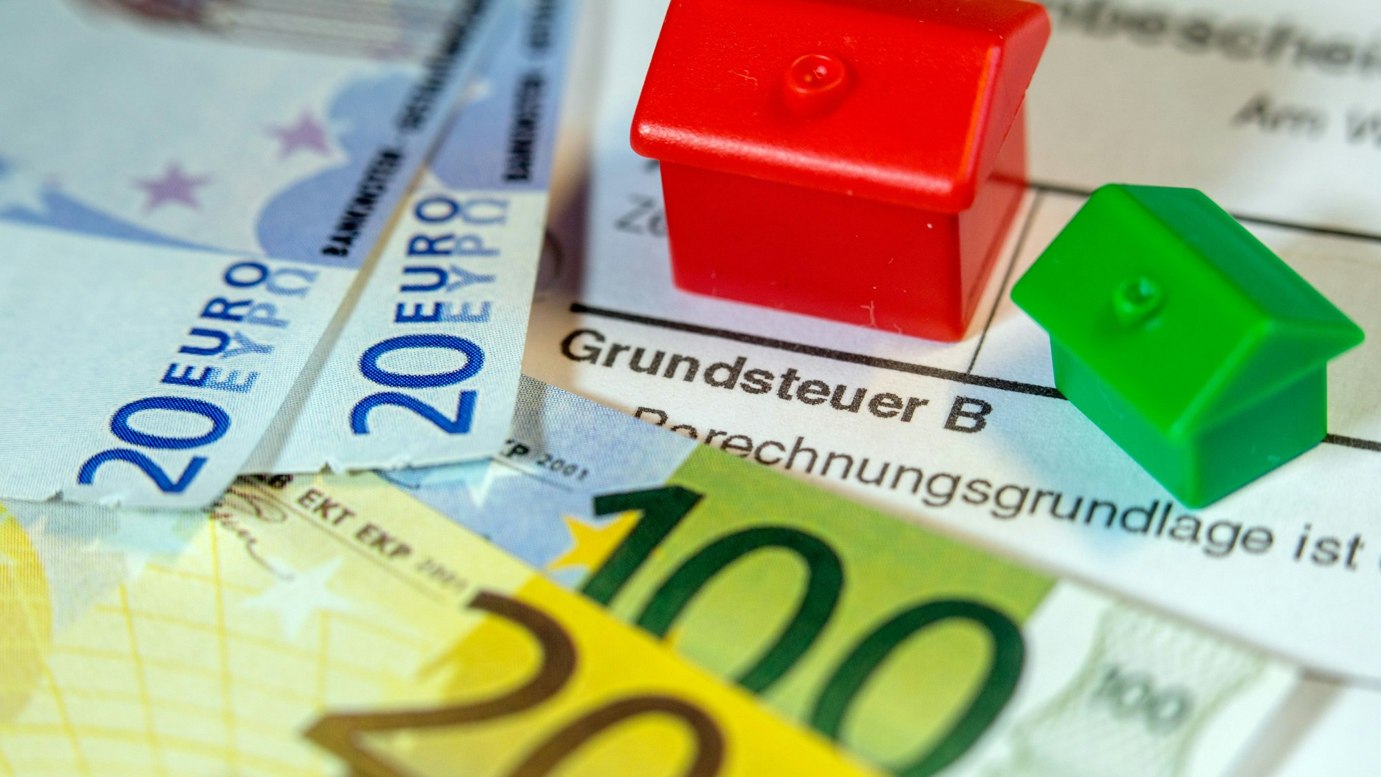 Zwei Spielzeughäuser und die Nachbildung von Euro-Geldscheinen liegen auf einem Abgabenbescheid für die Entrichtung der Grundsteuer.