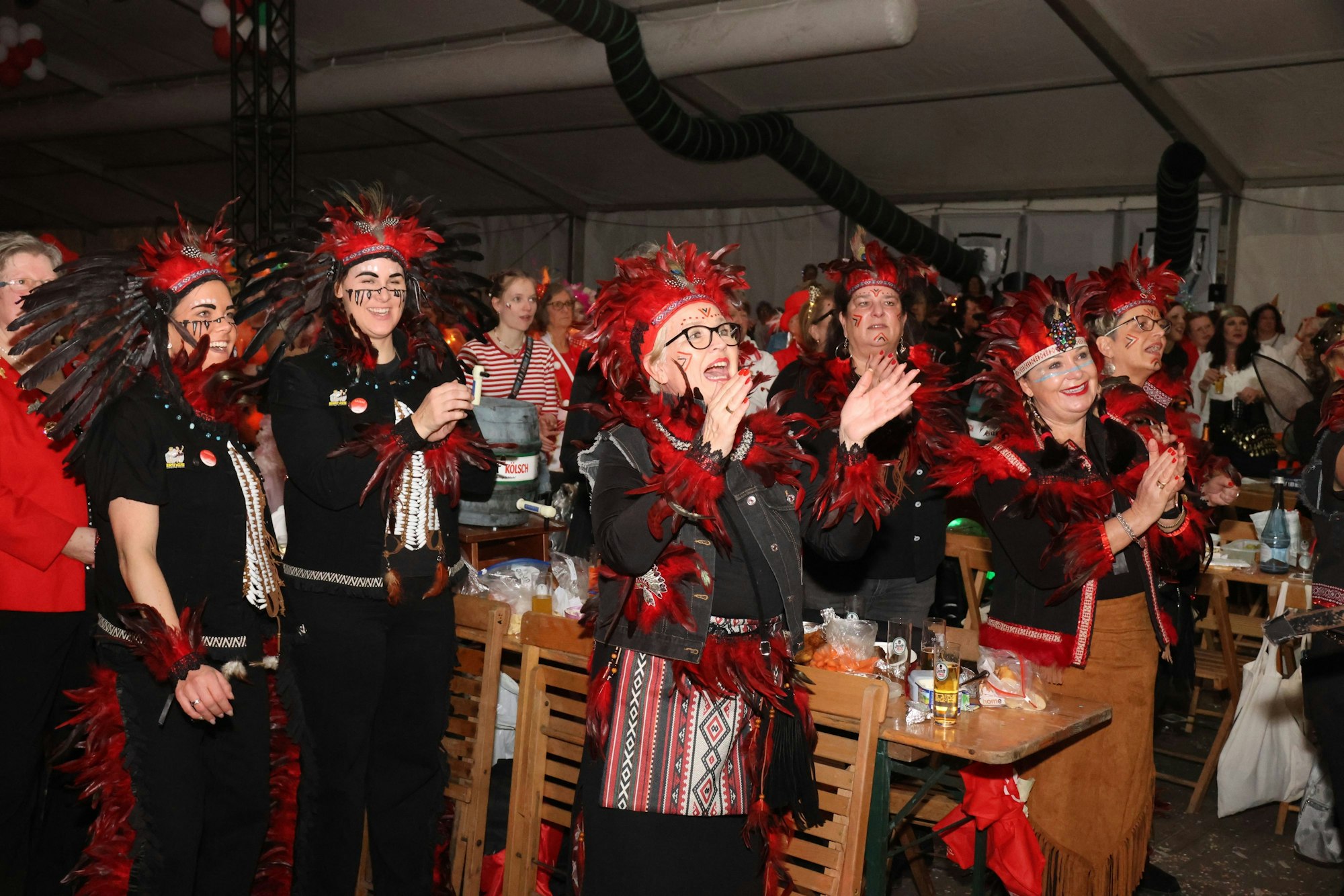 Eine Gruppe Frauen ist mit schwarz-roten Federn kostümiert. Die Frauenm lachen und klatschen.