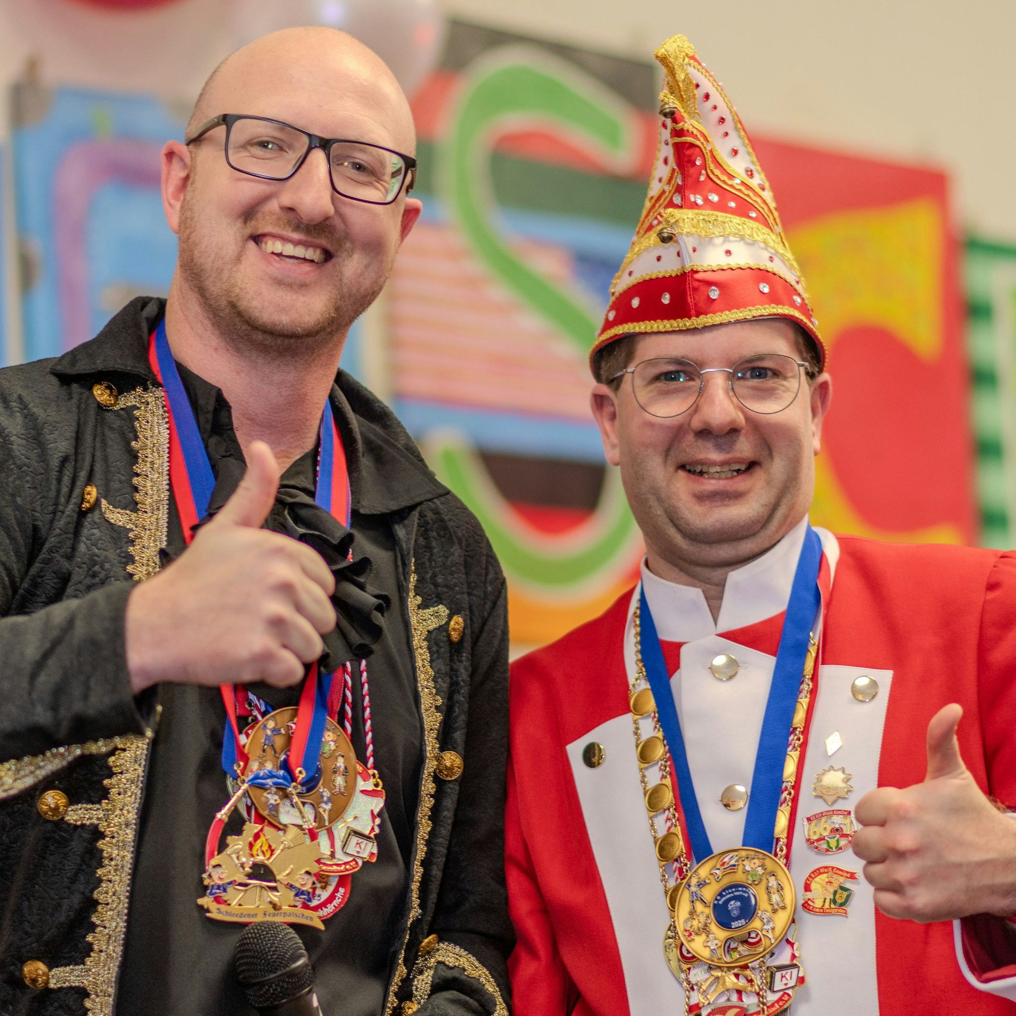 Ingo Pfennings und René Gerhards tragen mehrere Karnevalsorden um den Hals und lächeln in die Kamera.
