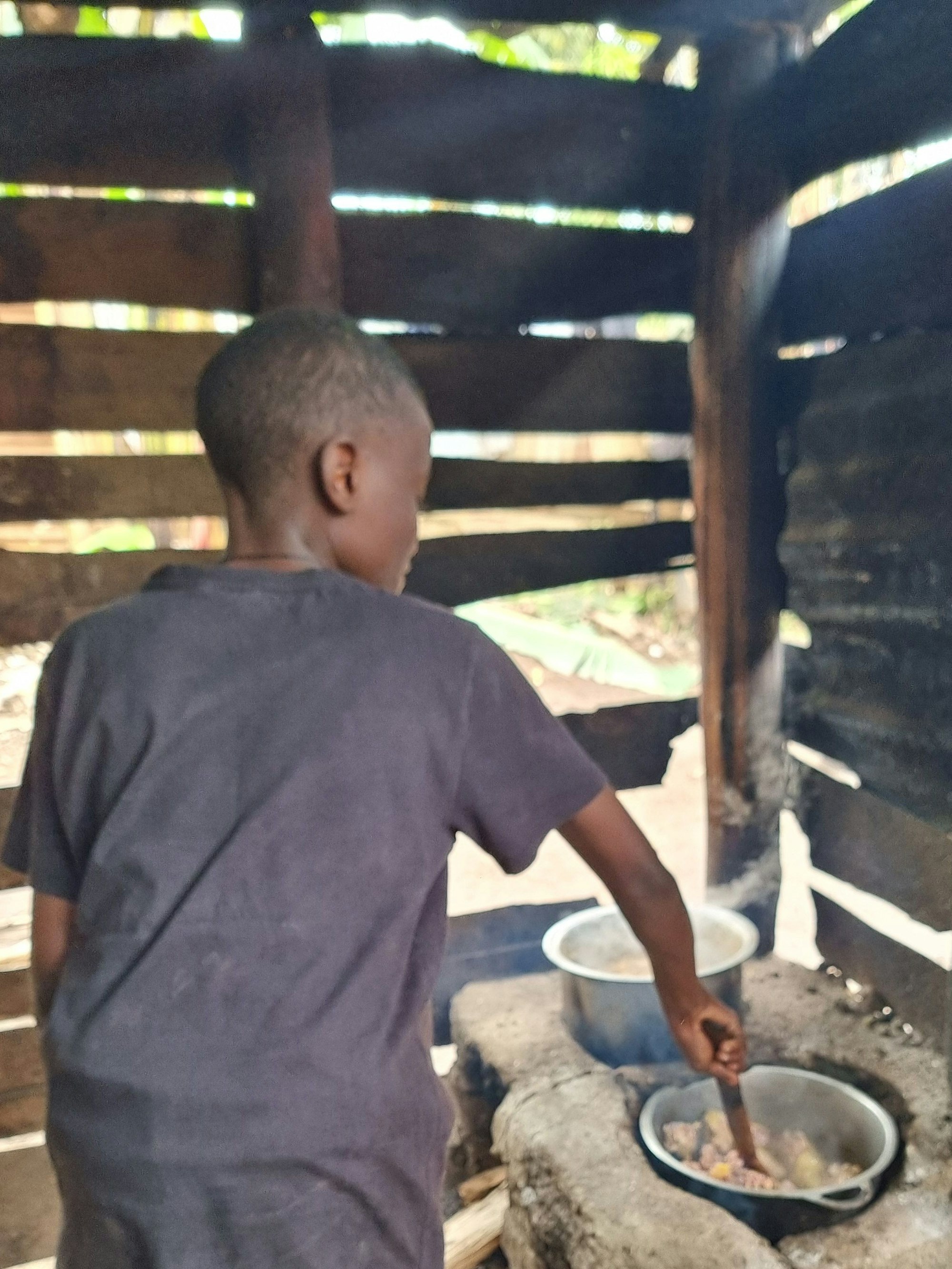 Ein Junge rührt ein Gericht in einem Topf um, das in einem Lehmöfchen warm gemacht wird.