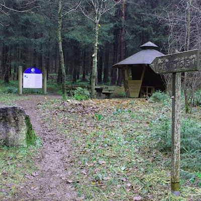 An einem historischen Stein im Wald stehen eine Infotafel und eine Schutzhütte.