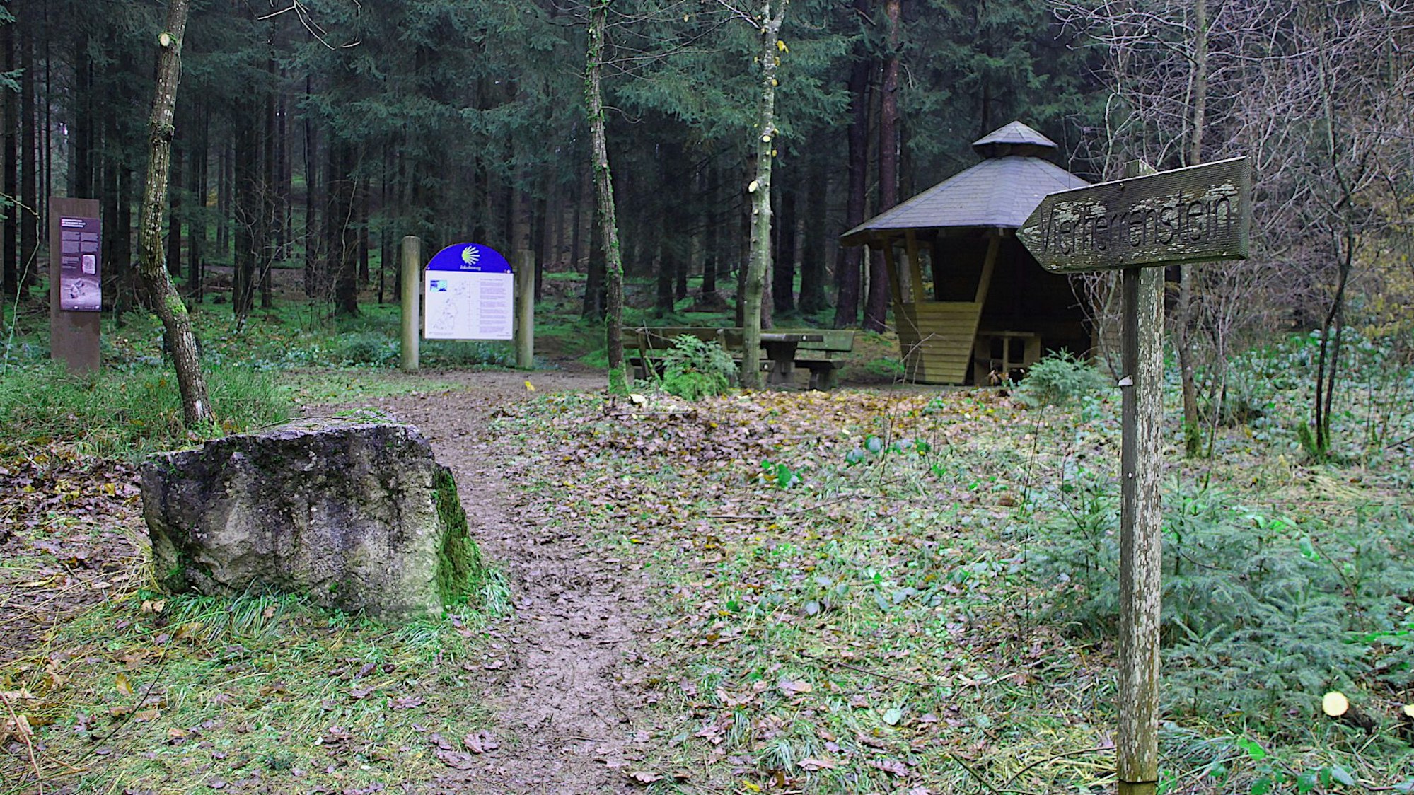 An einem historischen Stein im Wald stehen eine Infotafel und eine Schutzhütte.