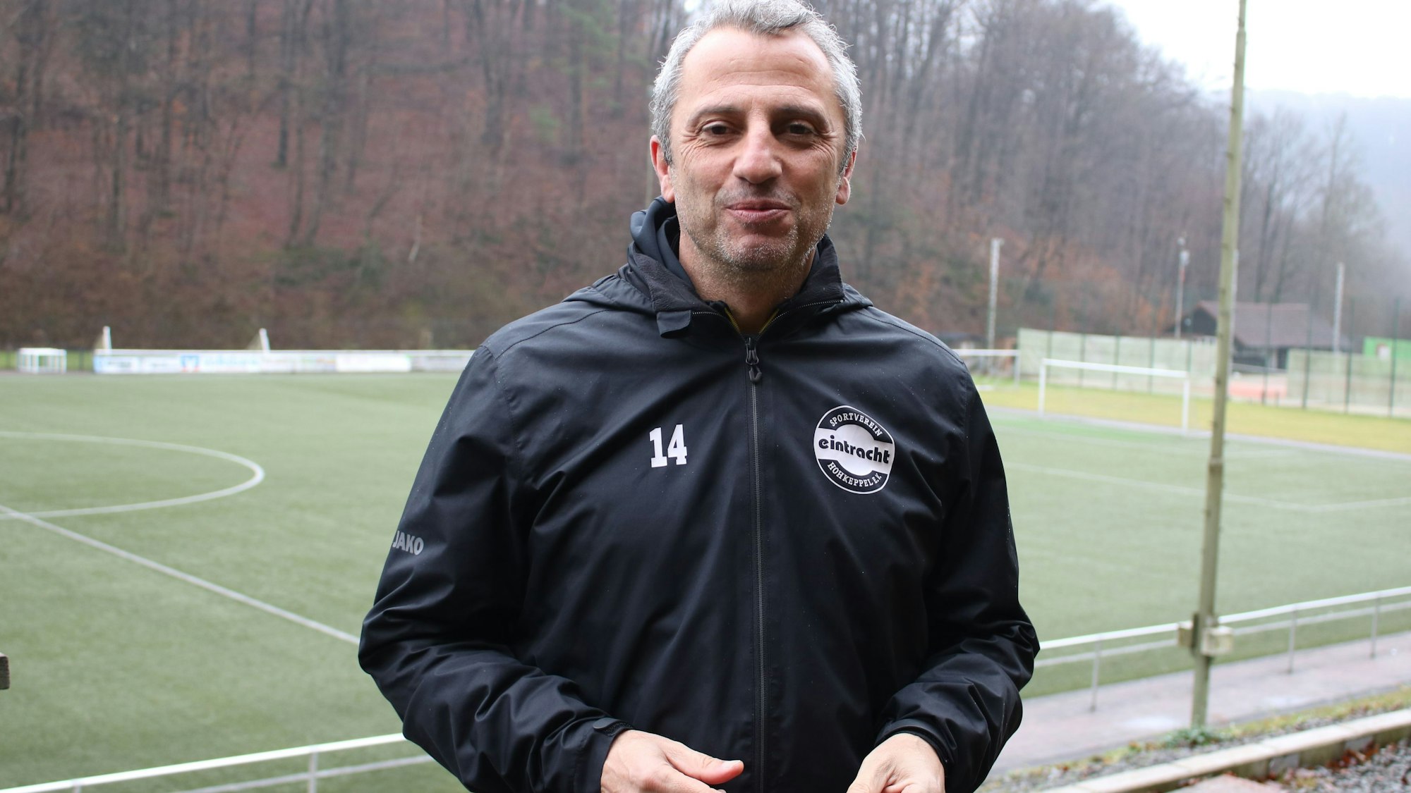 Ein Fußballtrainer steht auf einer Tribüne, im Hintergrund ist ein Fußballplatz zu sehen.