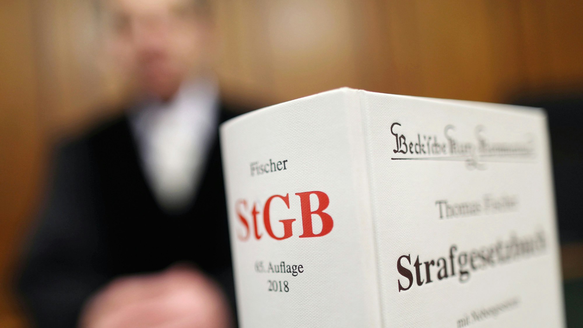 Das Symbolbild zeigt ein Strafgesetzbuch und im Hintergrund unscharf einen Menschen in einer Robe.