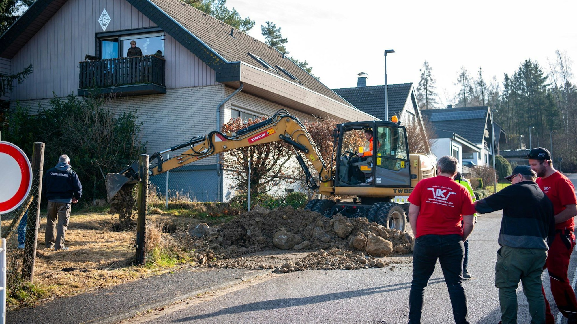 Ein Bagger, schaufelt Geröll auf eine Wiese, davor stehen in etwas Abstand ein paar Menschen.